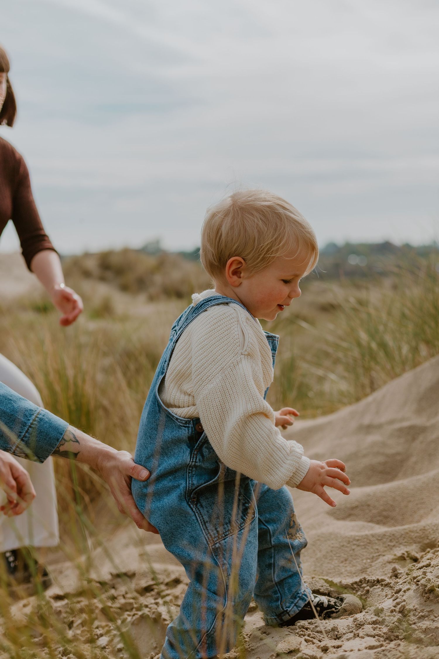 Toddler in denim overalls and white sweater playing on sandy dunes while being guided by an adult.