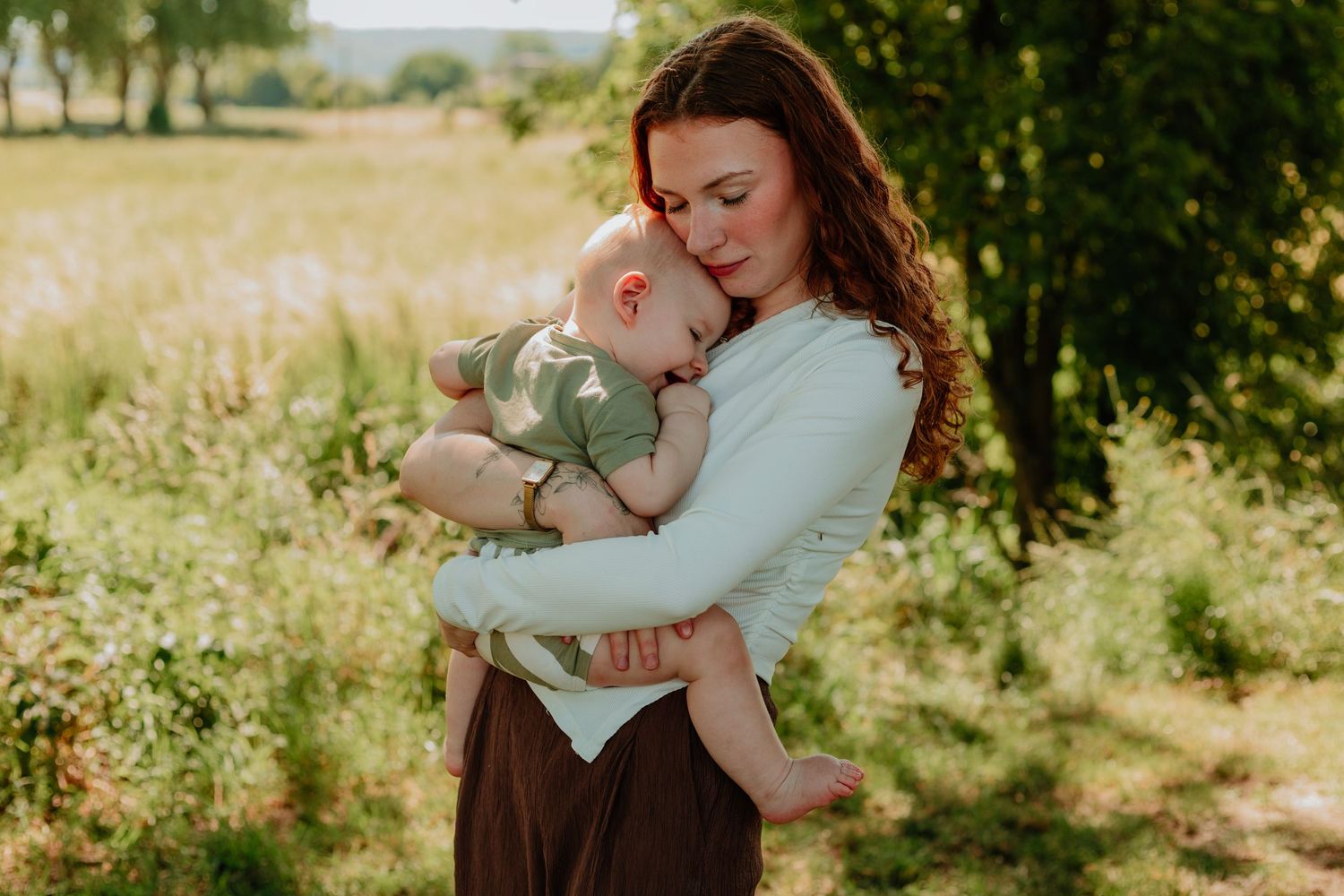 A mother tenderly holds her sleeping baby outdoors in a sunlit green field, wearing a soft blue top and brown skirt.