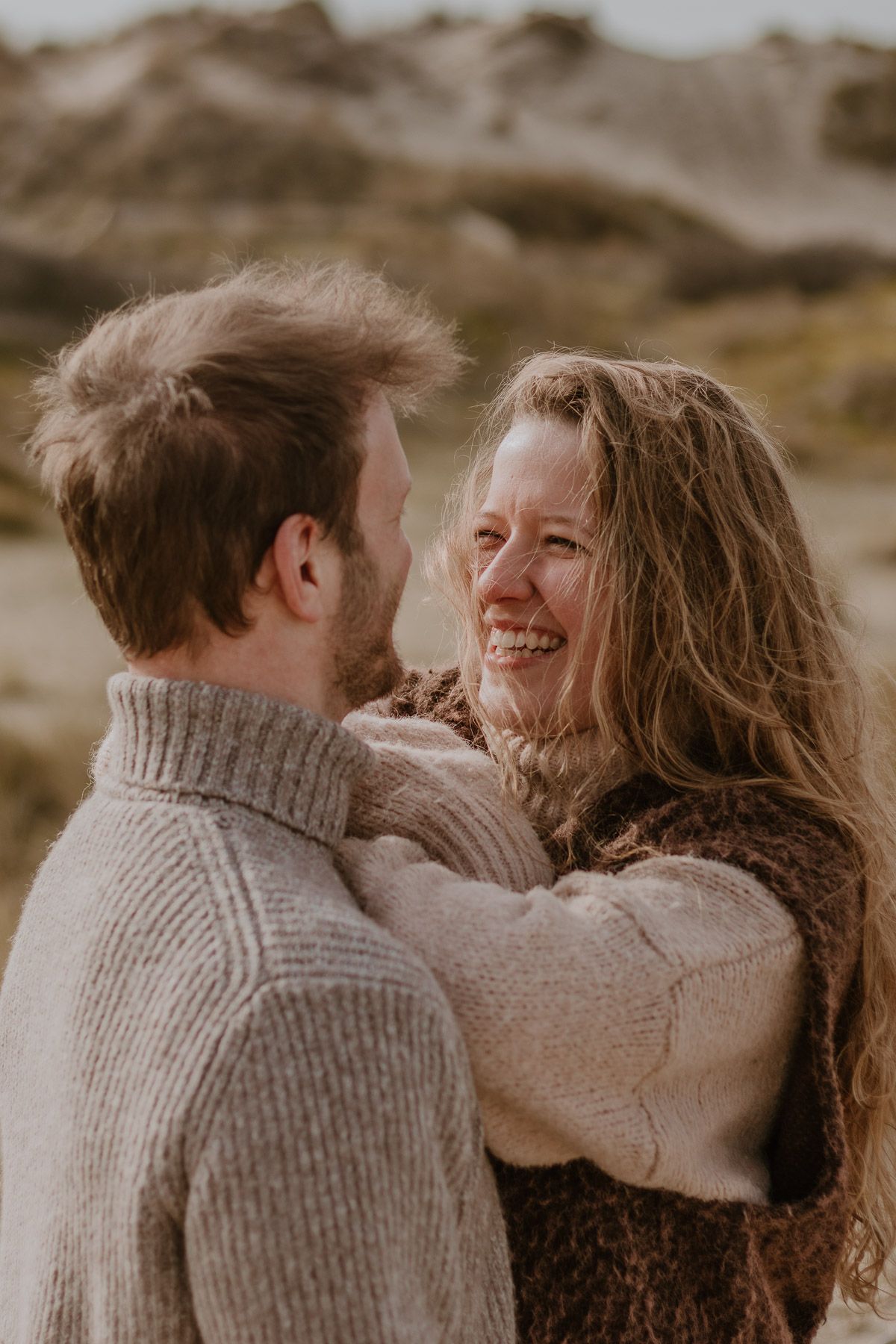 A smiling couple in cozy knit sweaters embrace outdoors, with windswept hair and sandy dunes in the background.