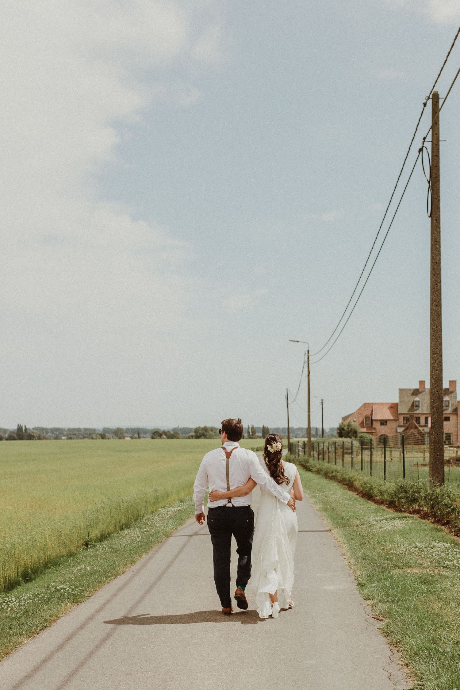 A couple walks down a rural dirt road lined with utility poles and green fields on a cloudy day.
