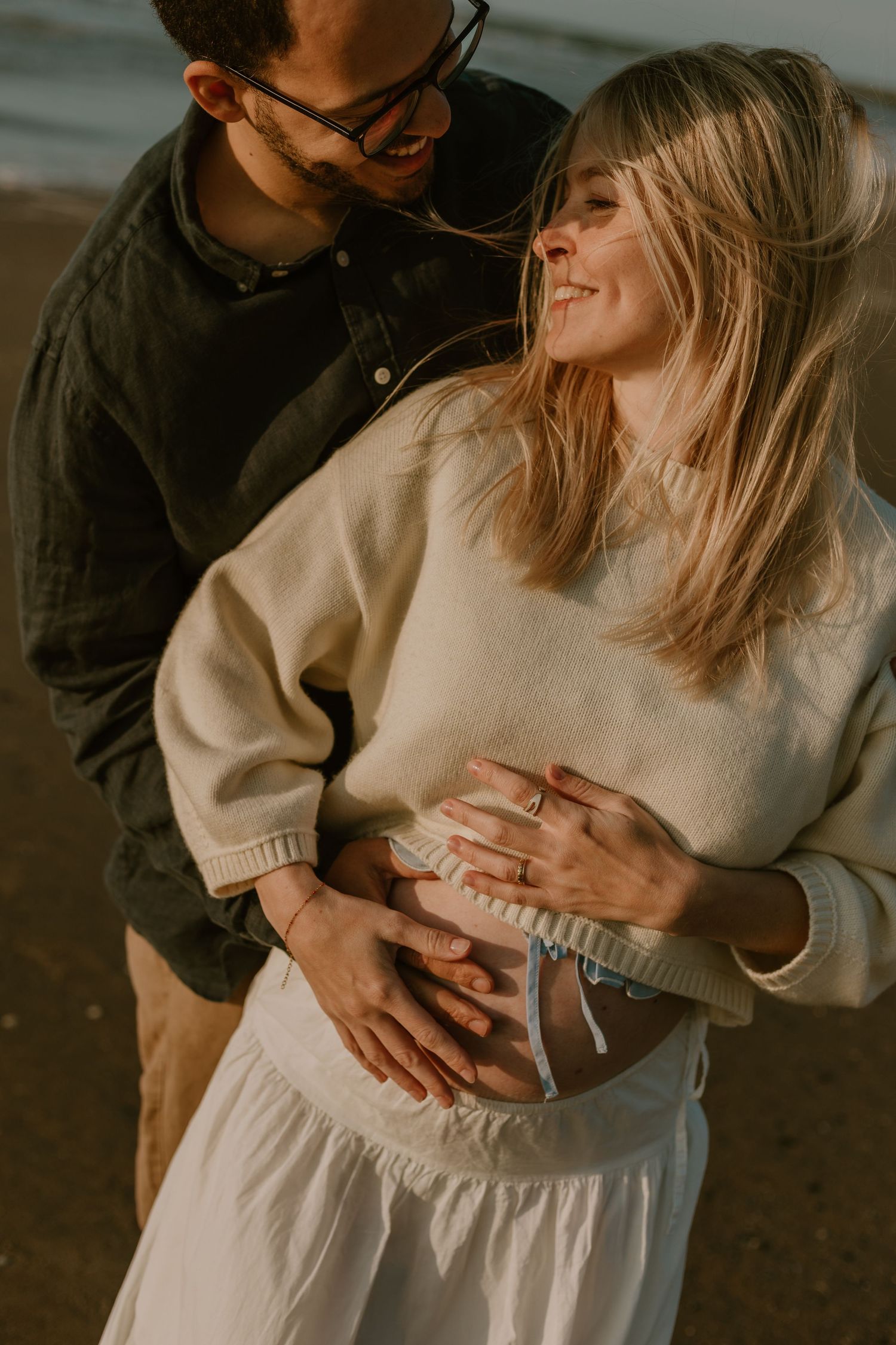 Couple embracing on beach, woman smiling with baby bump, wearing cream sweater and white skirt.