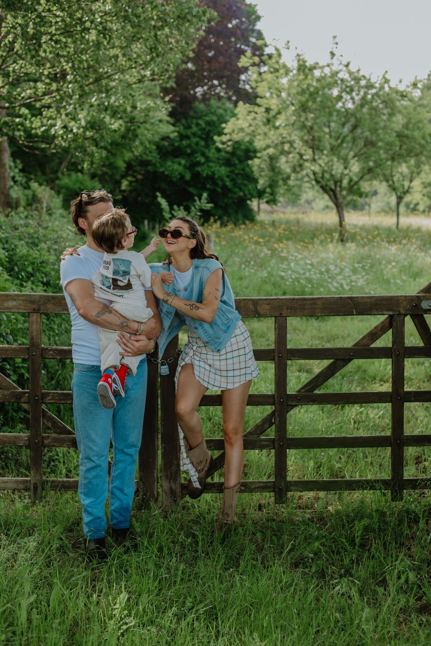 A young family with a toddler poses by a rustic wooden gate in a lush green countryside setting.