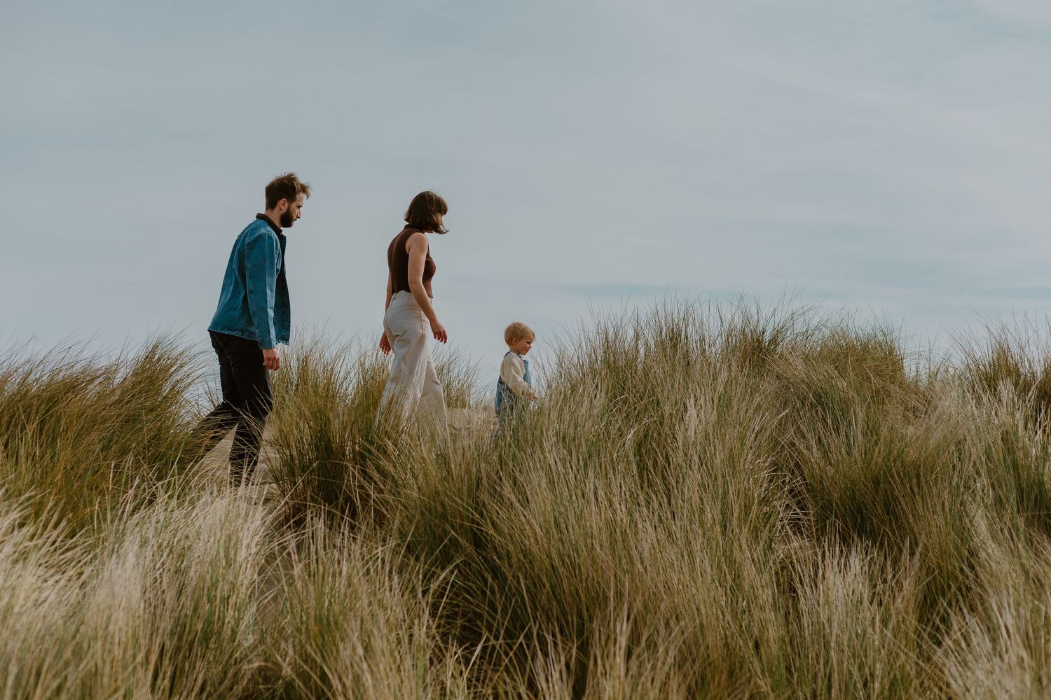 Family of three walking through tall grass on sand dunes under a cloudy sky.