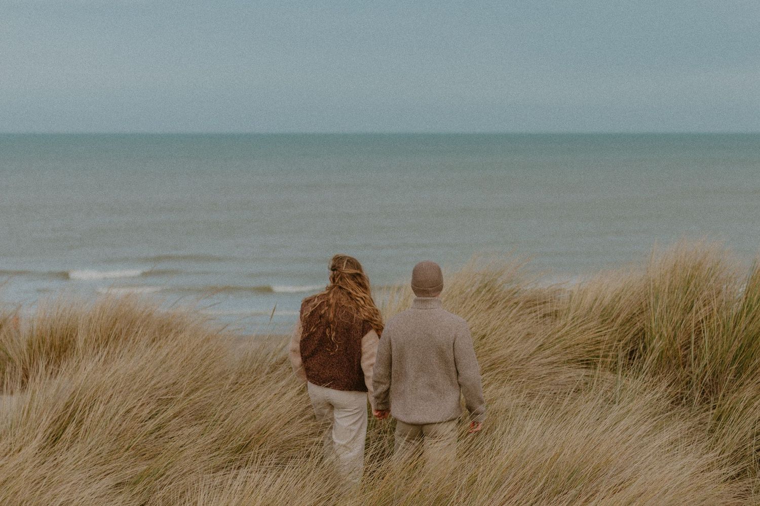 Two people stand together overlooking the ocean from grassy sand dunes on an overcast day.