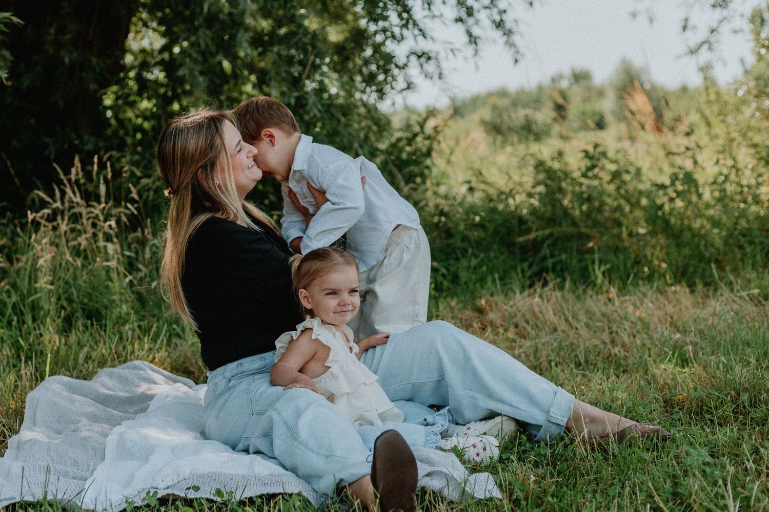 A mother sits on a blanket in a green meadow as her young son kisses her cheek and toddler daughter sits nearby.