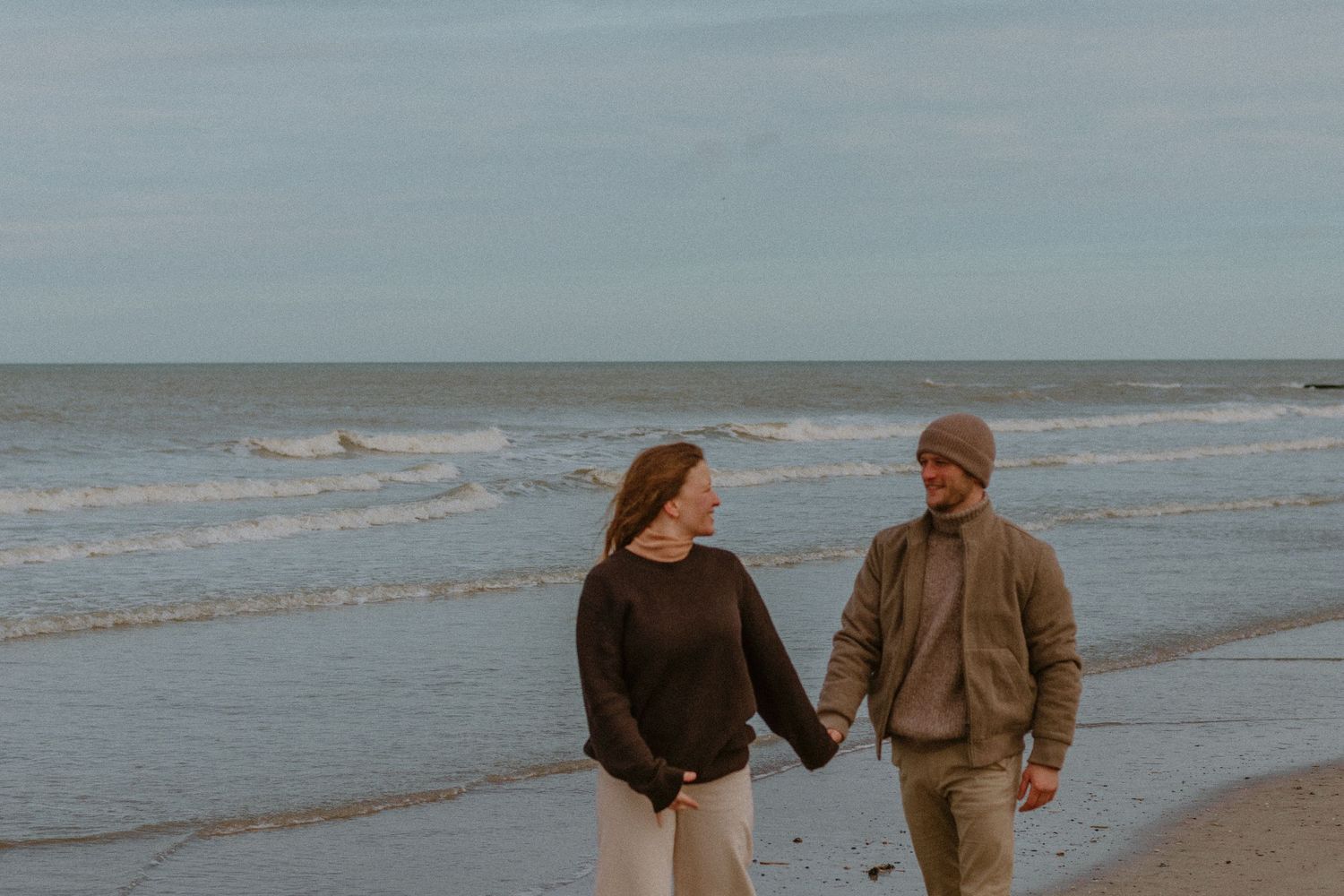 Two people walk along a misty beach shoreline on an overcast day with waves rolling in.