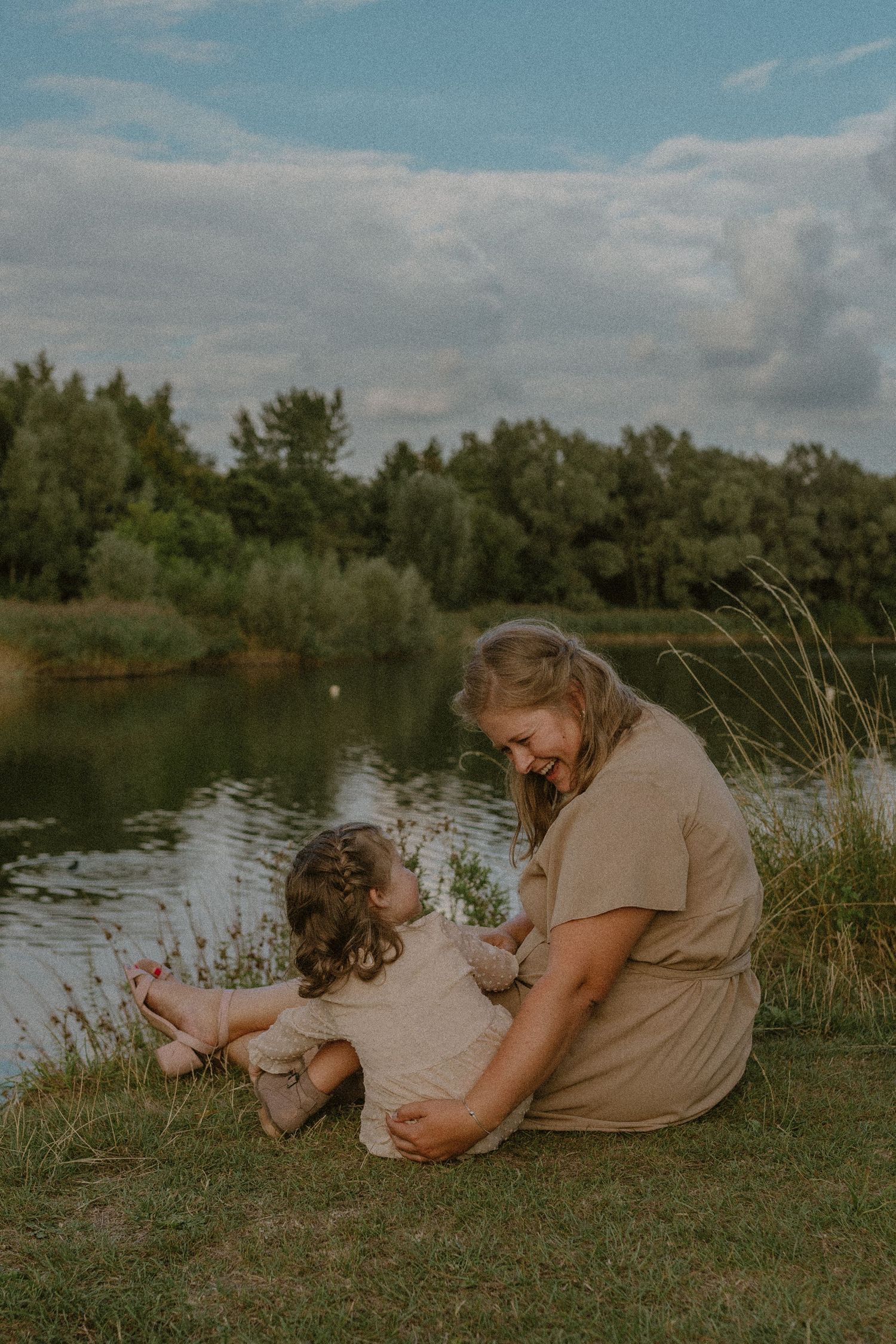Family sitting together on grassy riverbank during golden hour with calm water and trees in background.