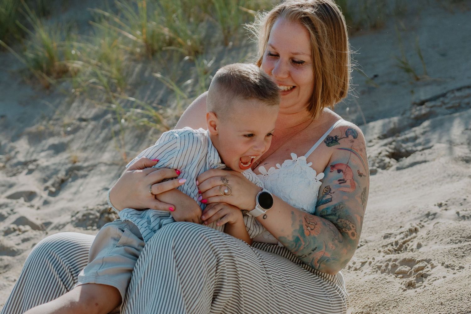 A mother with tattoos sits on a sandy beach, lovingly embracing her young toddler in a tender moment.
