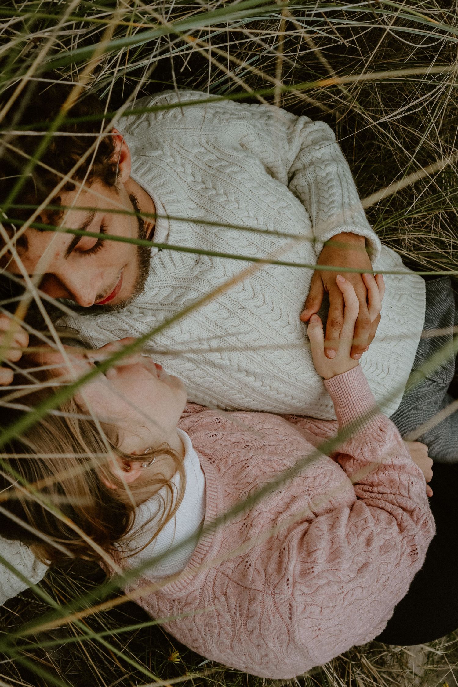 Een stel ligt in hoog gras, gekleed in witte en roze outfits, tijdens een romantische fotoshoot.