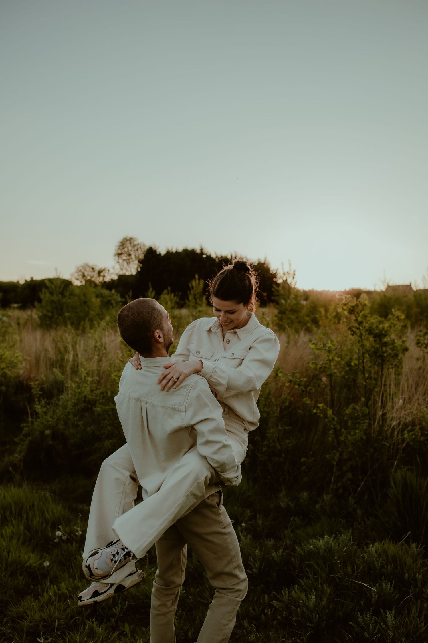 A romantic couple embraces in a field at sunset while wearing light colored clothing and casual attire.