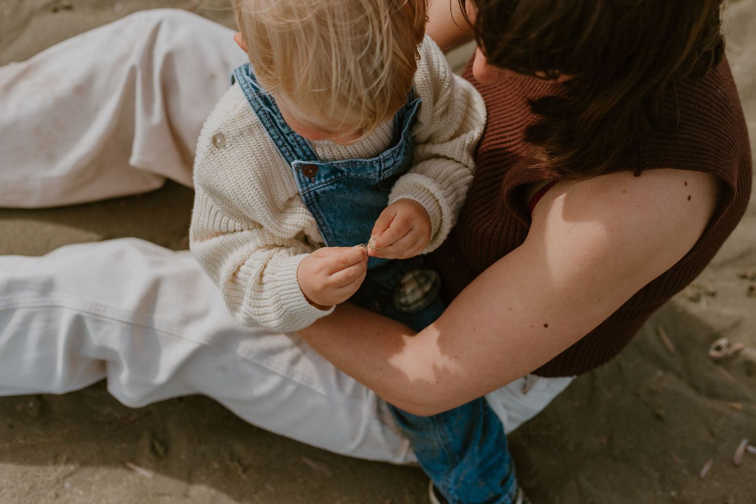 A mother sits on sandy beach holding her toddler in denim overalls, sharing a tender moment outdoors.