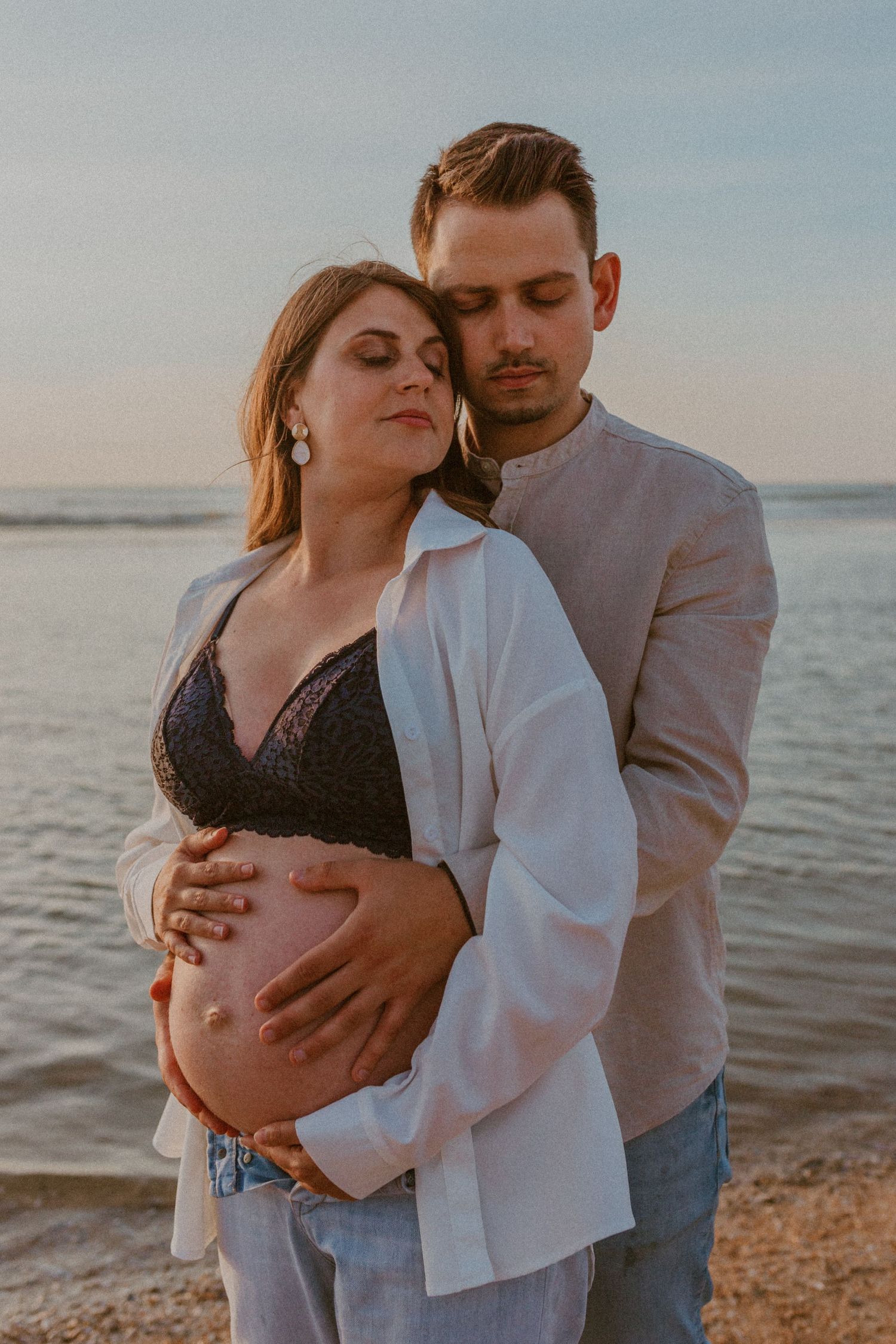 A couple embraces on a beach at sunset while showing off their baby bump in a maternity photoshoot.