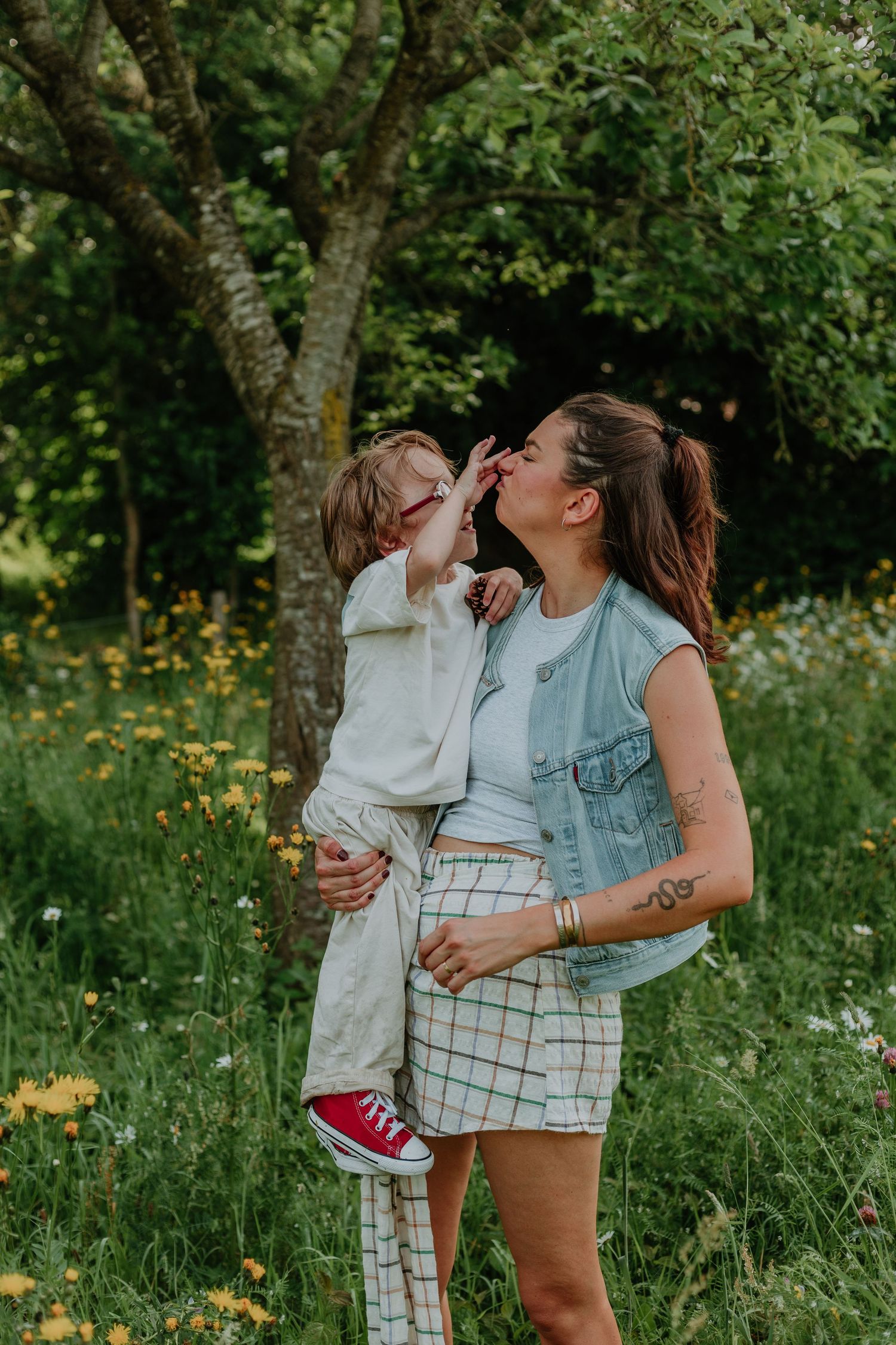 A woman holds a young child for a kiss in a lush green meadow with wildflowers and trees.