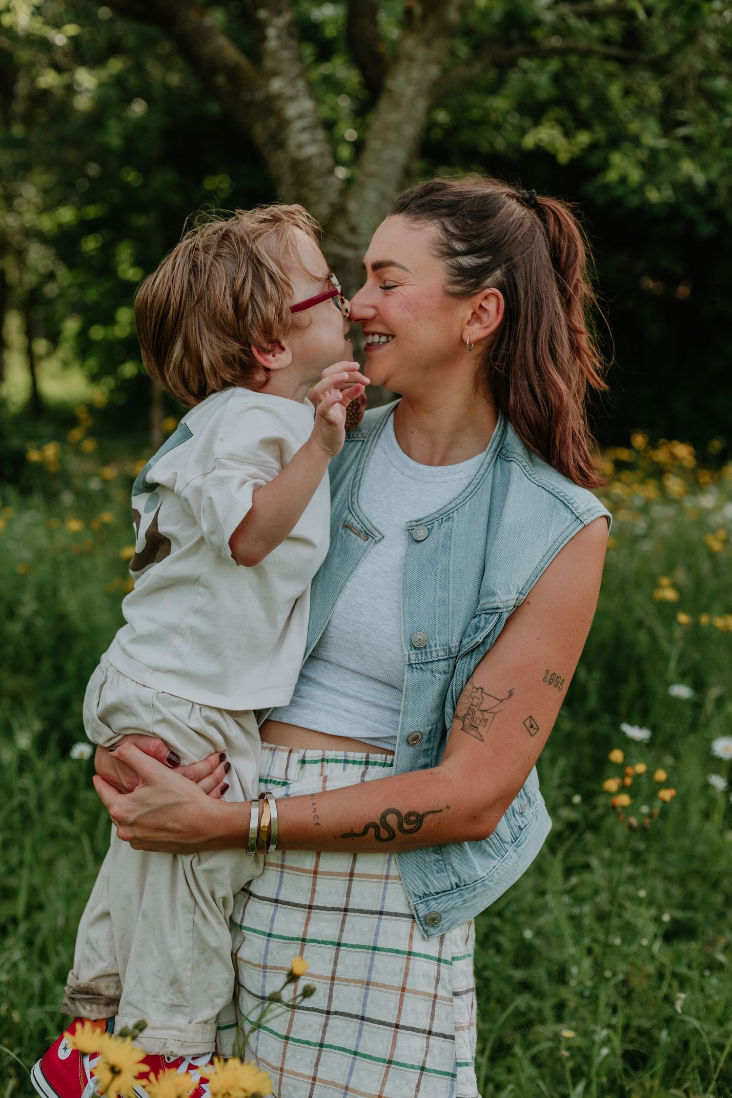 A woman in a denim vest holds a young child in a sunny garden, sharing a playful nose-to-nose moment.