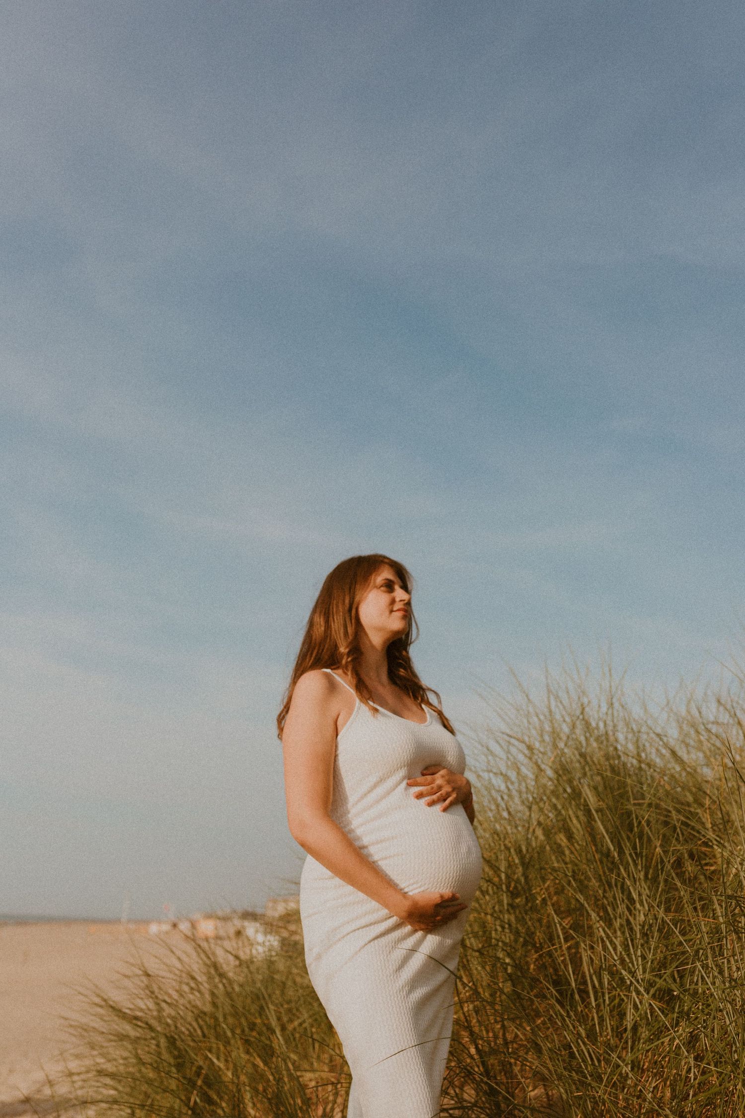 A pregnant woman in a white dress stands among beach grass dunes against a soft blue sky.