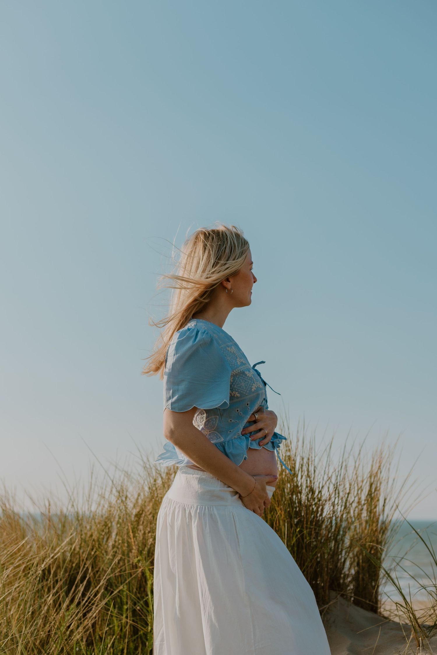 Pregnant woman in blue crop top and white skirt holding baby bump on sandy beach dunes under clear sky.