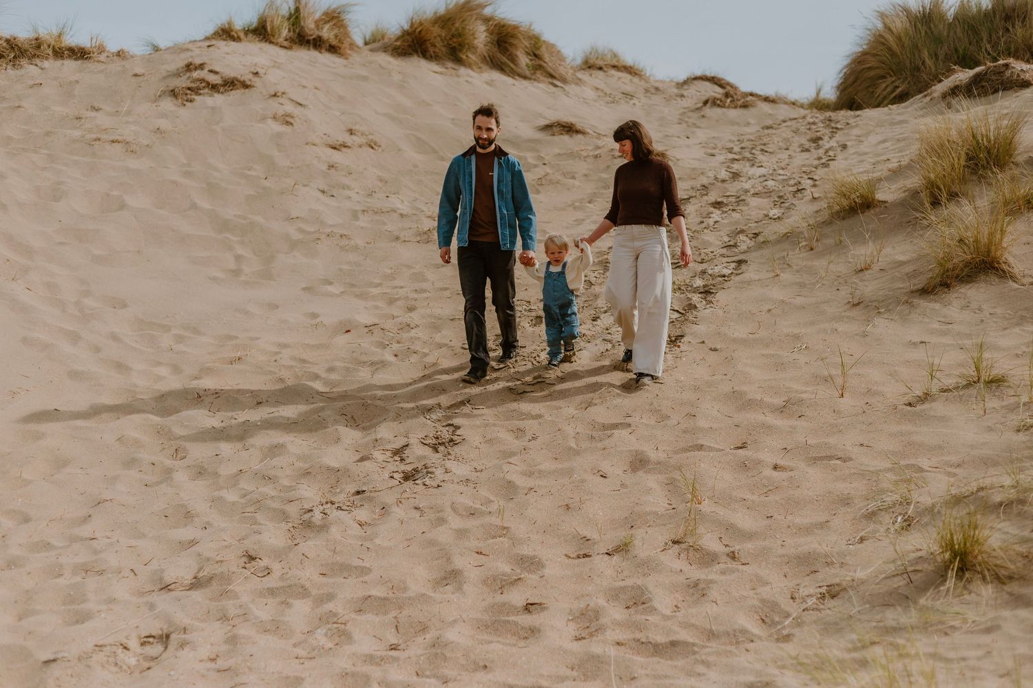 Family of three walking on sandy beach dunes, parents holding toddler's hands, wearing casual clothes on a cloudy day.