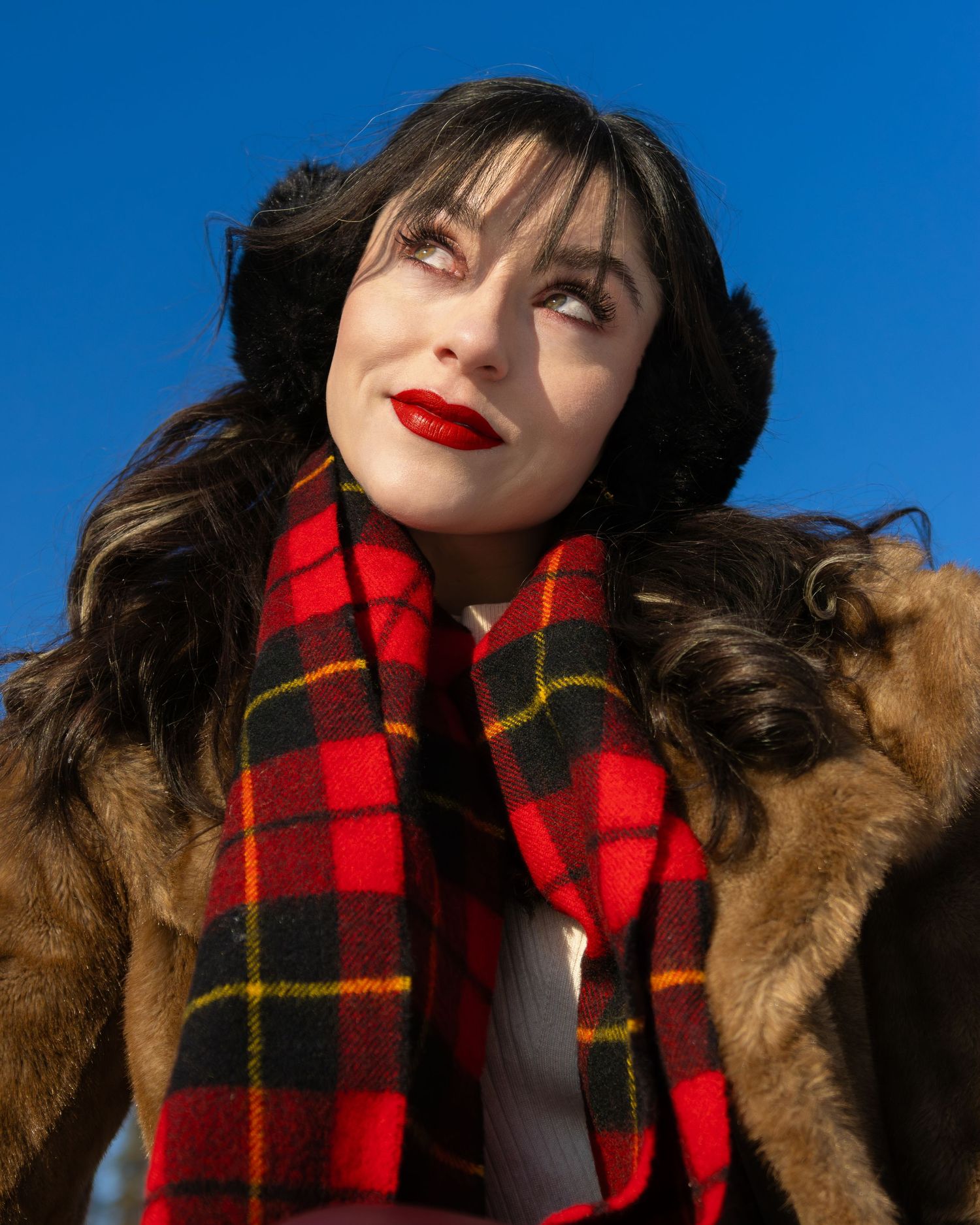 Woman with red lips and dark hair wearing a red plaid scarf and fur coat against a blue sky.