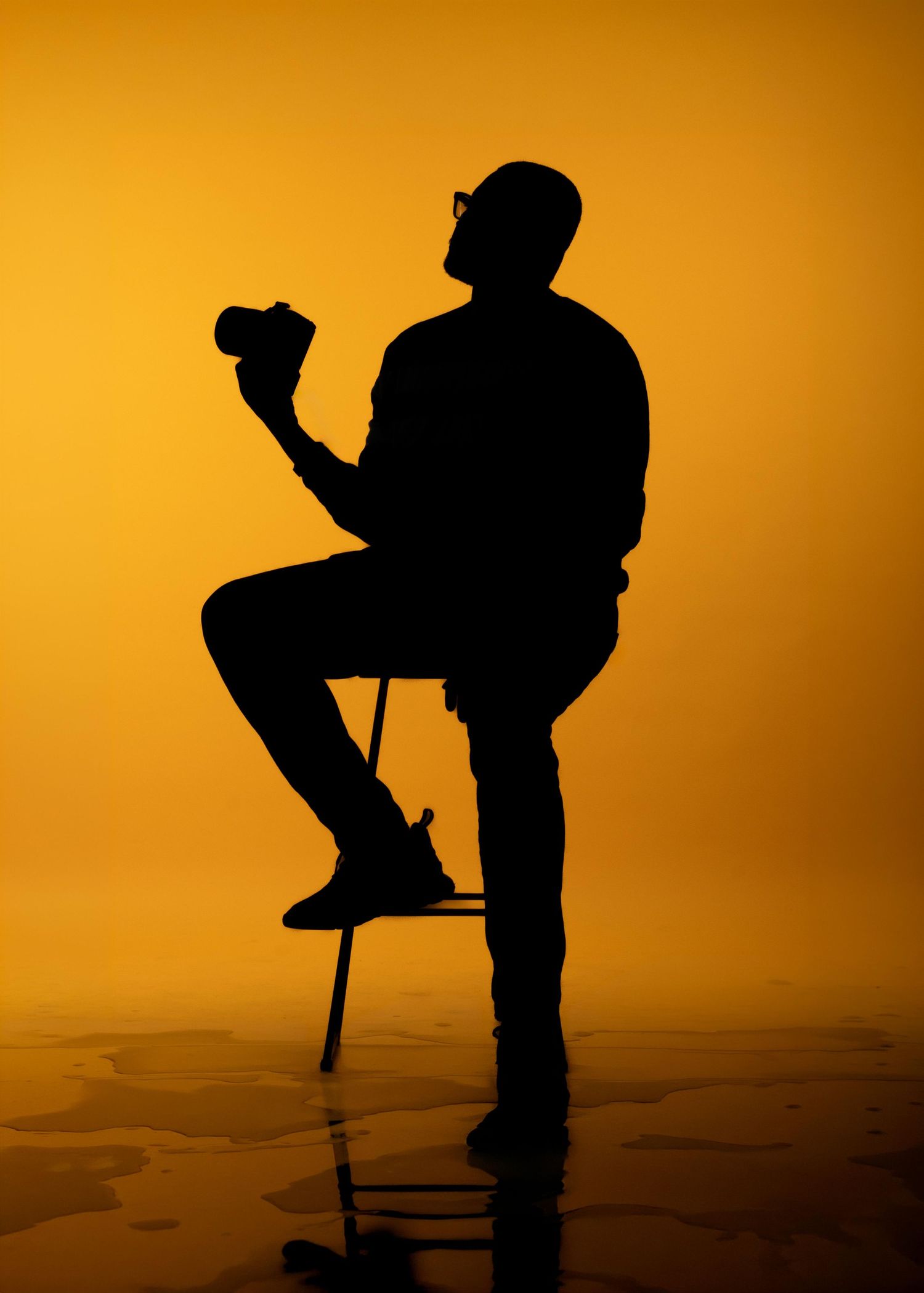 Silhouette of a man sitting on a stool holding a camera against a vibrant orange background.
