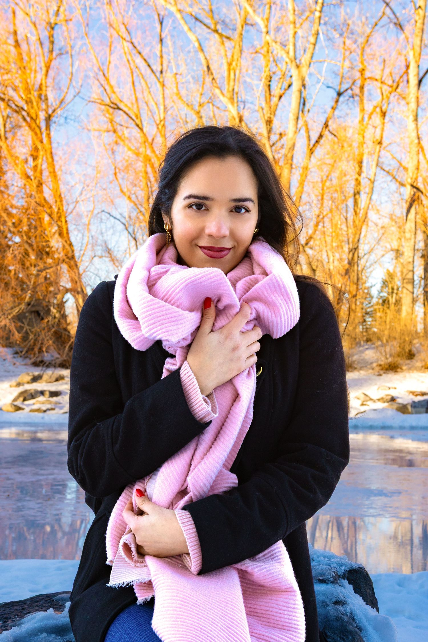 Young woman in black coat and pink scarf smiling outdoors in winter, with snow and golden trees in background.