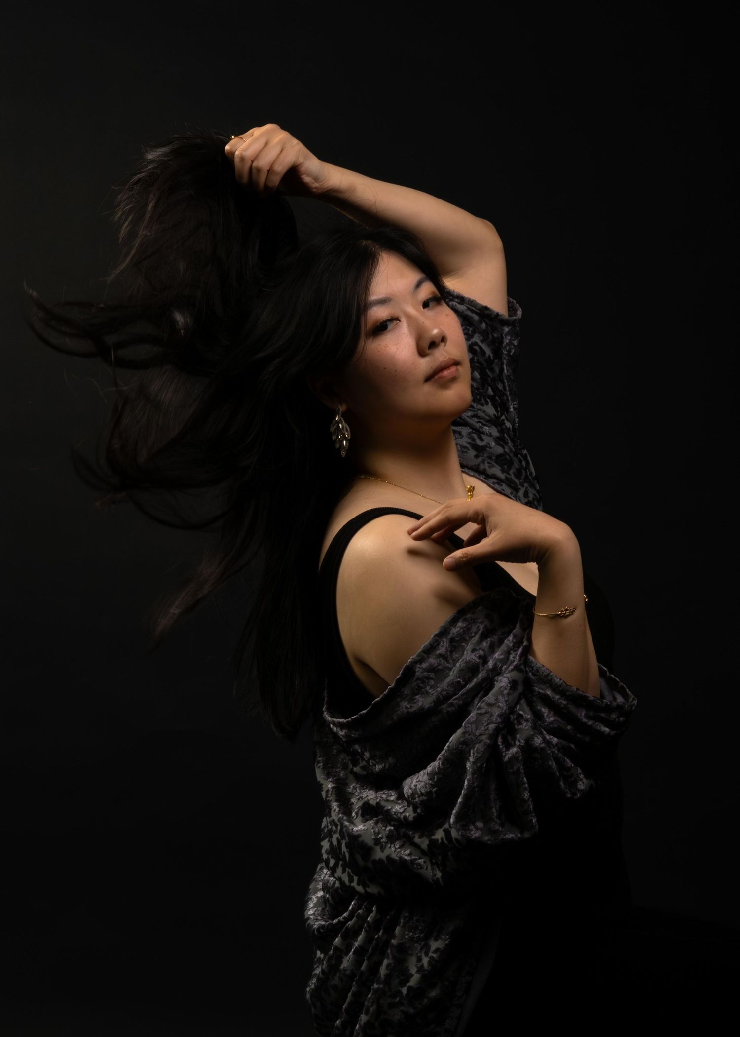 Woman in black dress poses dramatically with flowing hair against dark background, wearing elegant earrings.