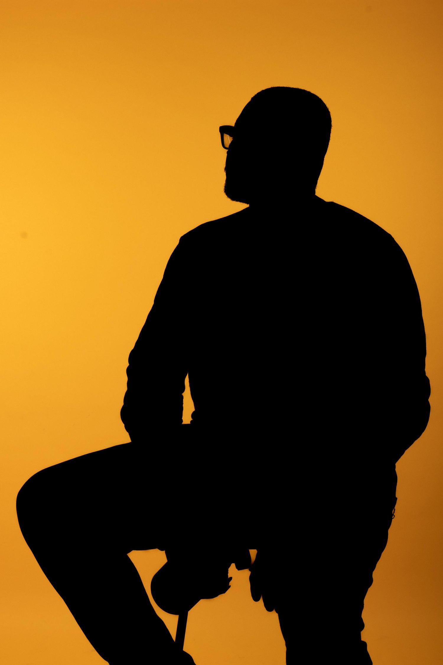 Silhouette of a man wearing glasses and a cap, sitting on a stool against a vibrant orange background.