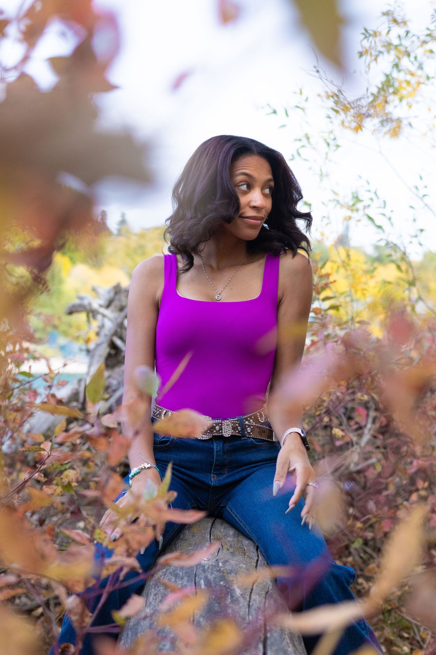 Woman in purple tank top and jeans sitting on a log surrounded by colorful autumn foliage outdoors.
