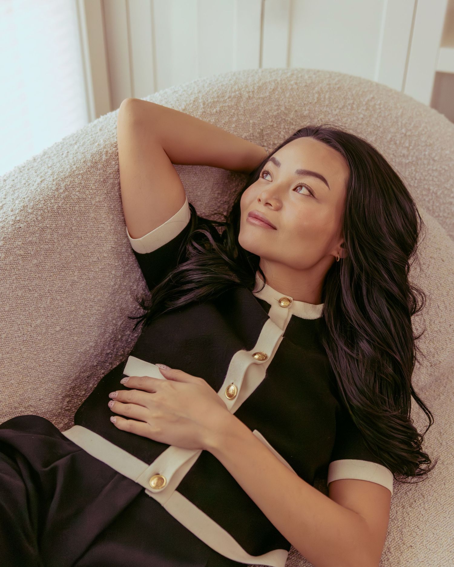 Woman with long dark hair reclines in a plush chair wearing a black and white dress with gold buttons.