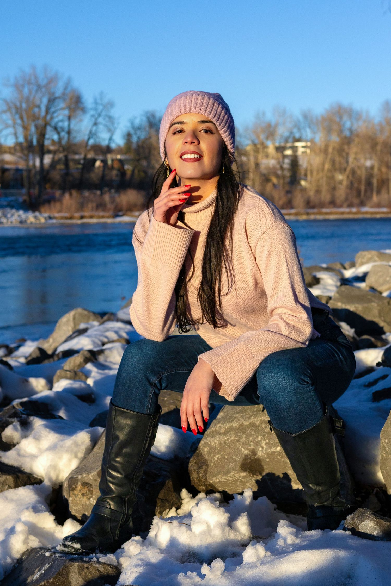 Woman in pink sweater and beanie sits on snowy rocks by a river in winter sunlight, wearing black boots and jeans.