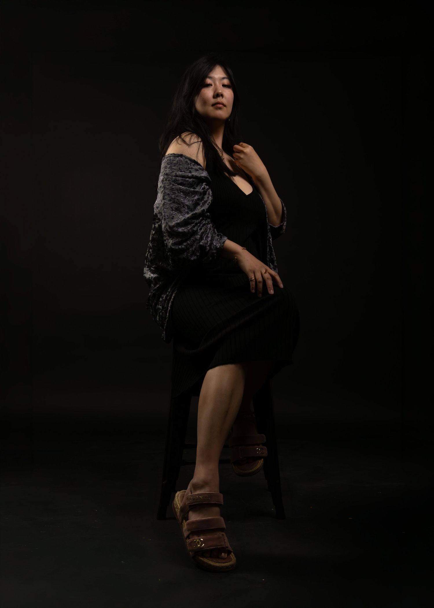 Woman in black dress and gray shawl poses elegantly on stool against dark background wearing brown sandals.