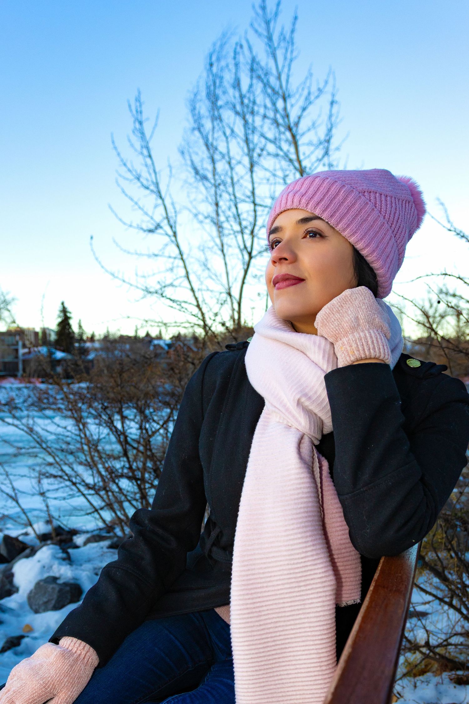 Woman in pink knit hat, scarf, and mittens sitting outdoors in a snowy winter landscape under a blue sky.