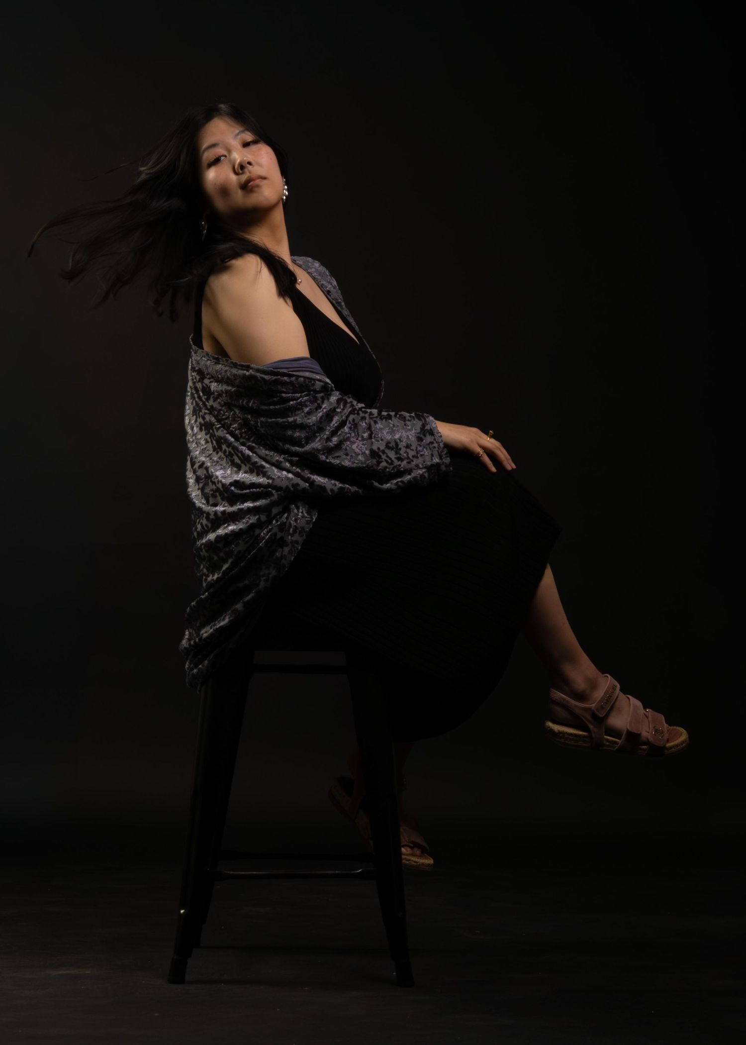 Woman in velvet kimono jacket poses dramatically on stool against dark background with wind-swept hair.