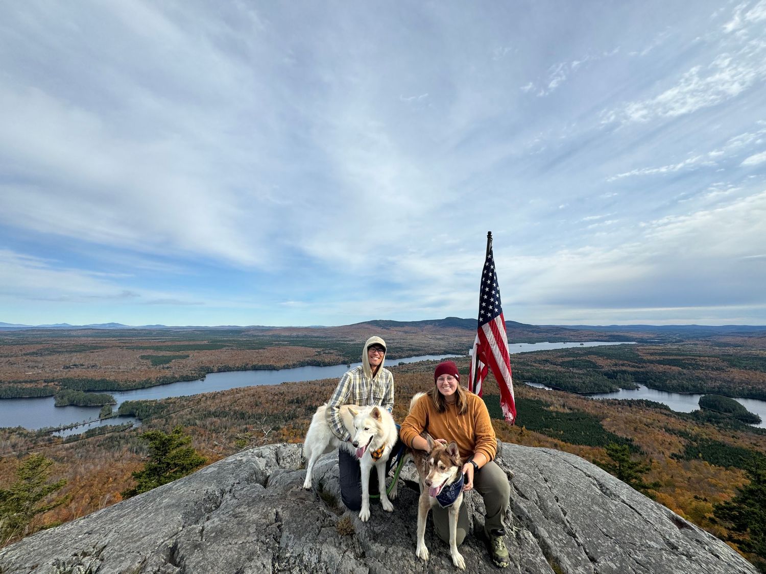 Man and woman with two huskies on top of Mosquito Mountain in The Forks, Maine during the fall with an American flag