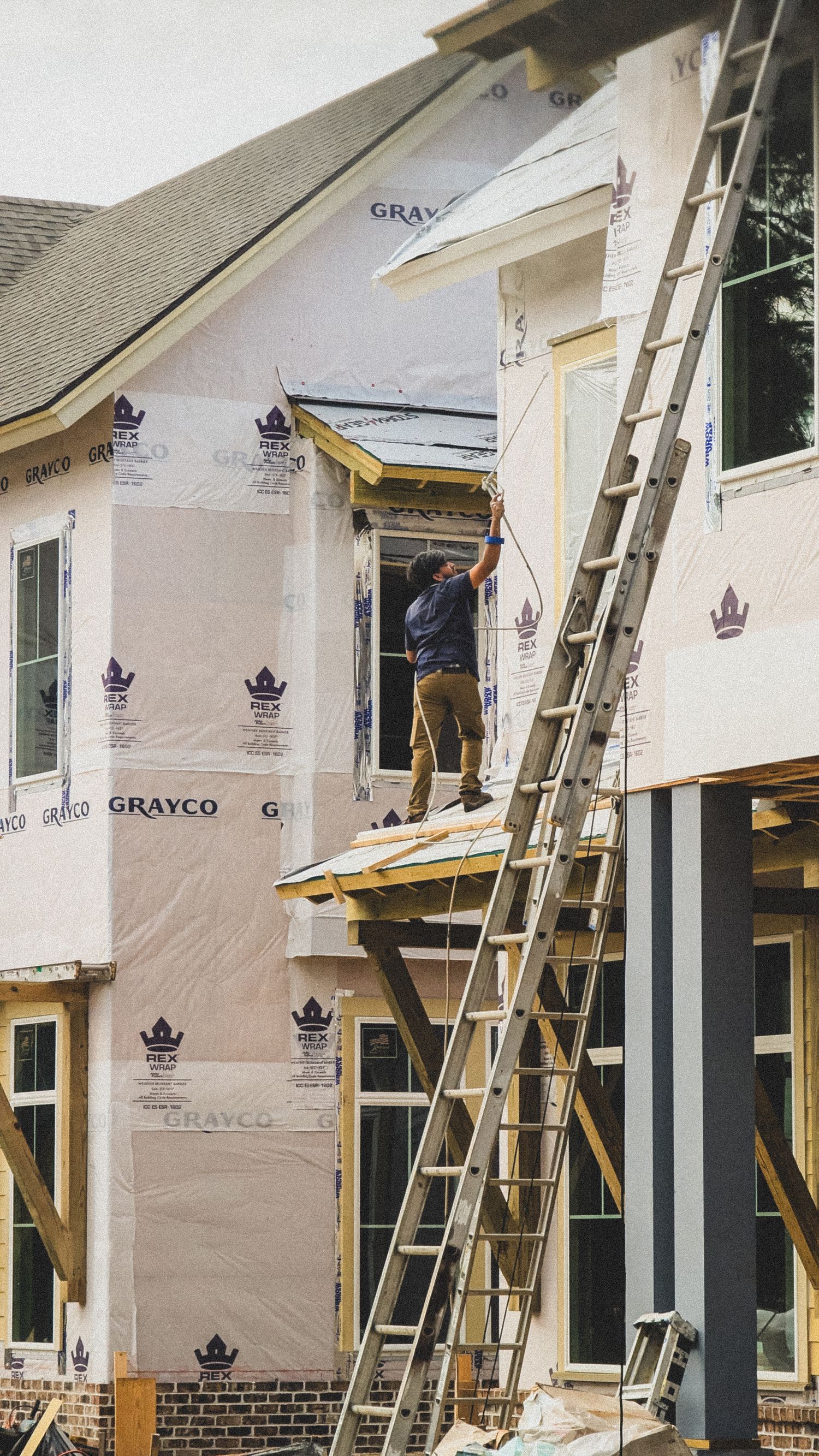 On-site video production of Construction workers install house siding and weatherproofing materials on a residential building exterior.