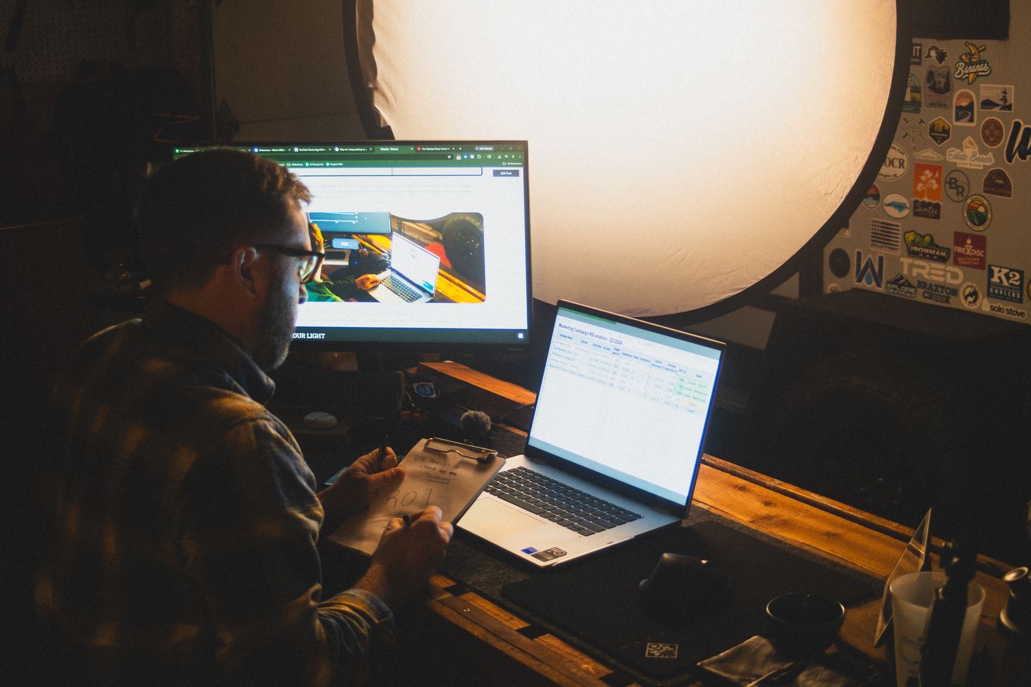 High-Res BTS Workshop Photo showing David working at desk with multiple laptop screens reviewing a Marketing ROI 2026 spreadsheet for