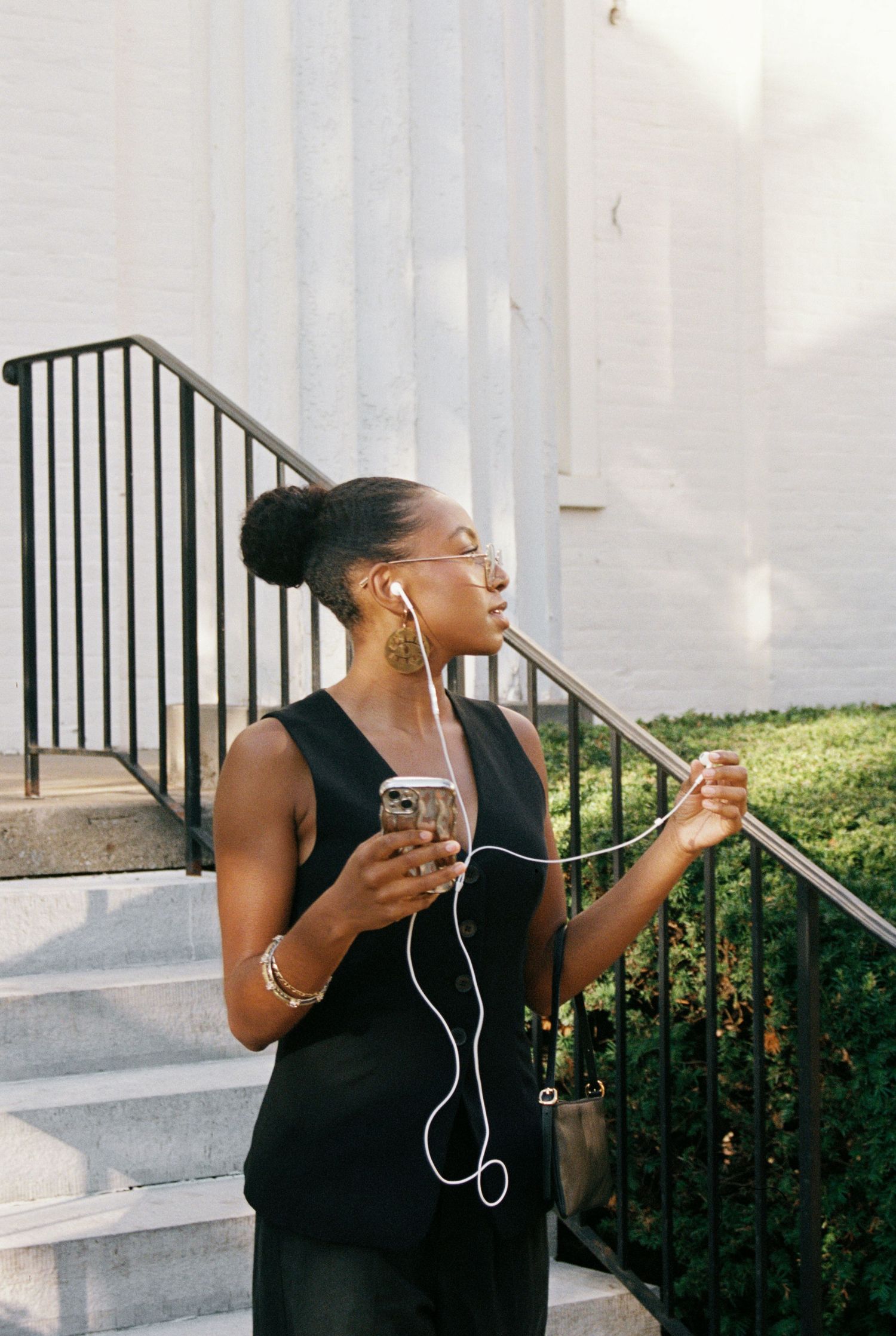 Casual moment on white steps with earphones and black dress against architectural details.