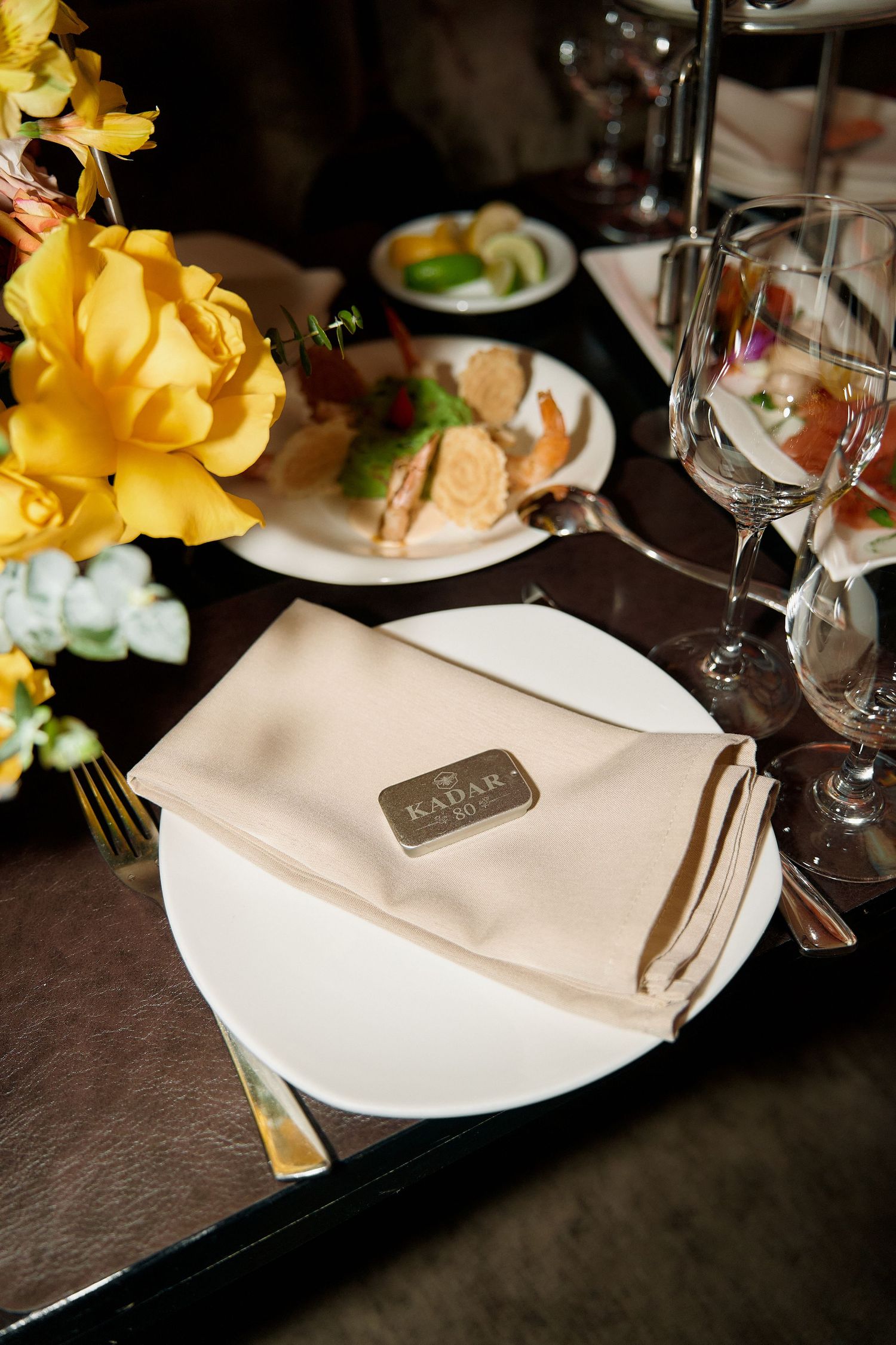 Elegant place setting with beige napkin and Kadar restaurant card on white plate with yellow roses nearby.
