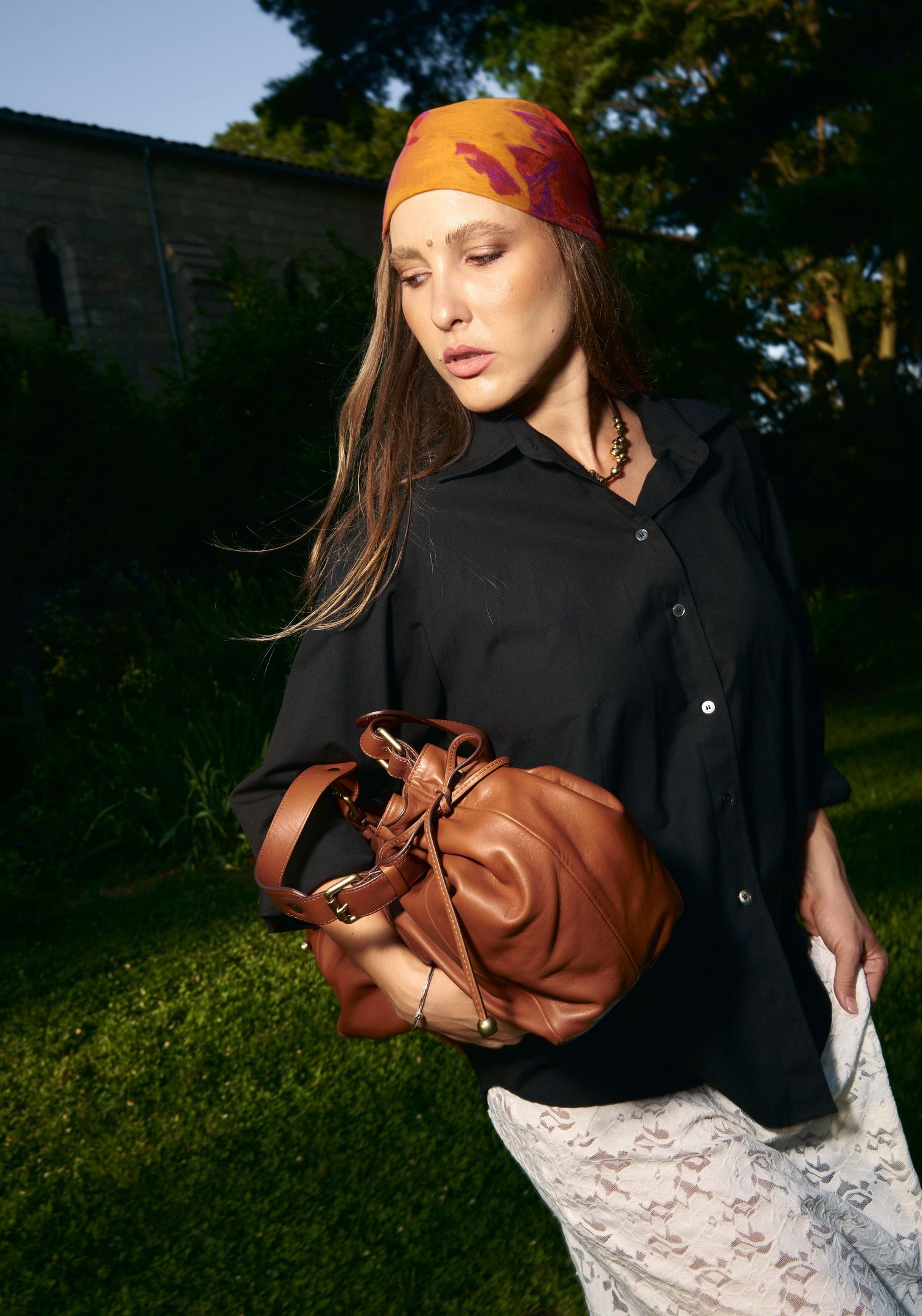 Woman wearing orange headscarf and black shirt holding brown leather bucket bag outdoors.