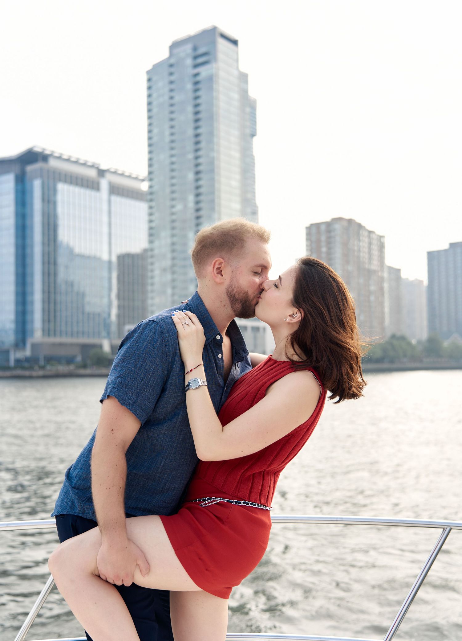 Couple kissing on yacht bow with city skyline and river in background.