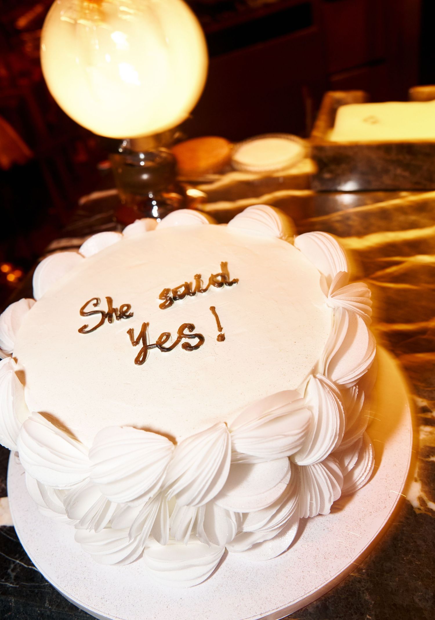White celebration cake with gold script reading She said yes on a decorative pedestal stand.
