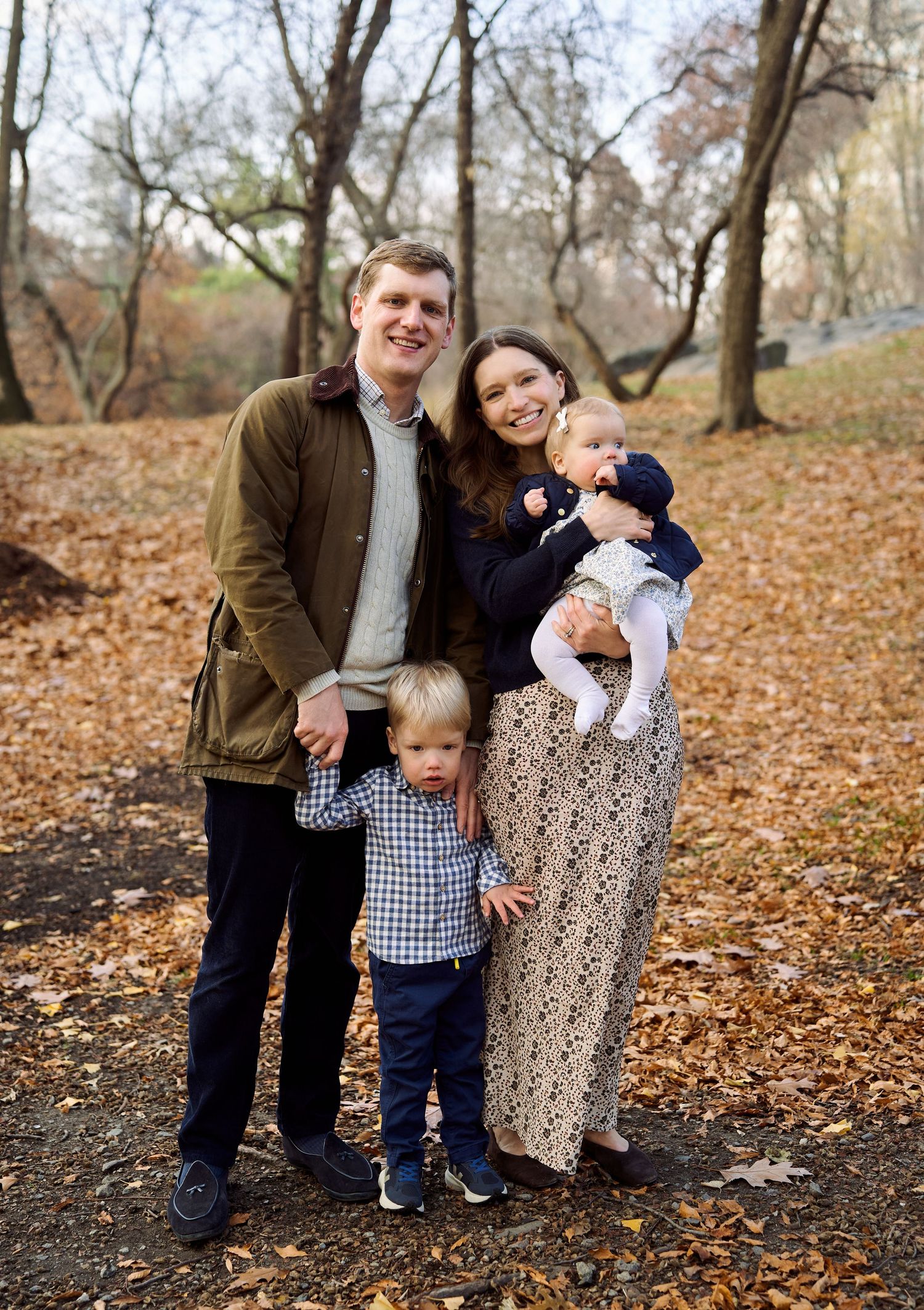 Family of four posing on a leaf-covered path in autumn park, parents with toddler and infant.