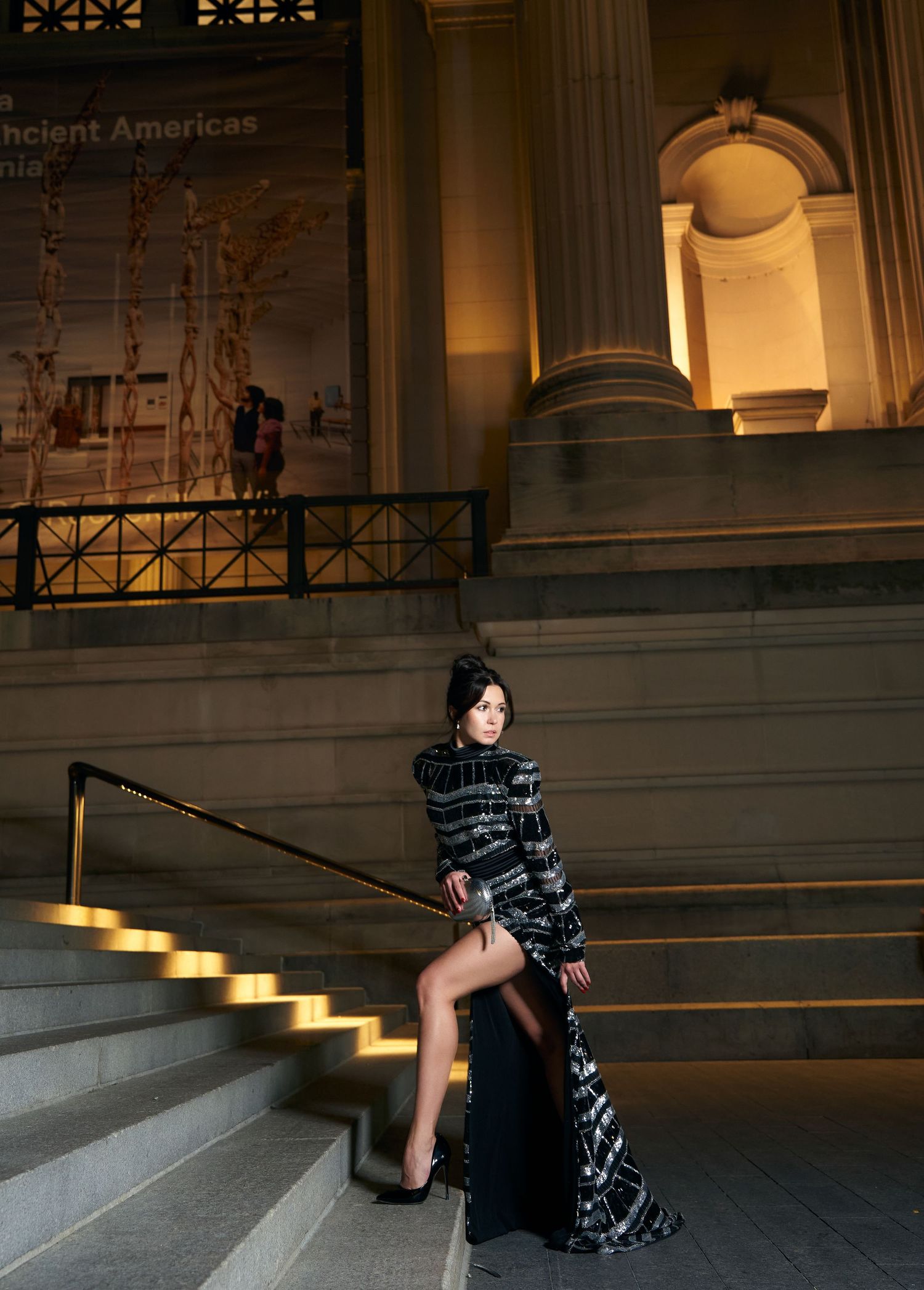 A woman in a black sequined gown poses on museum steps at night.