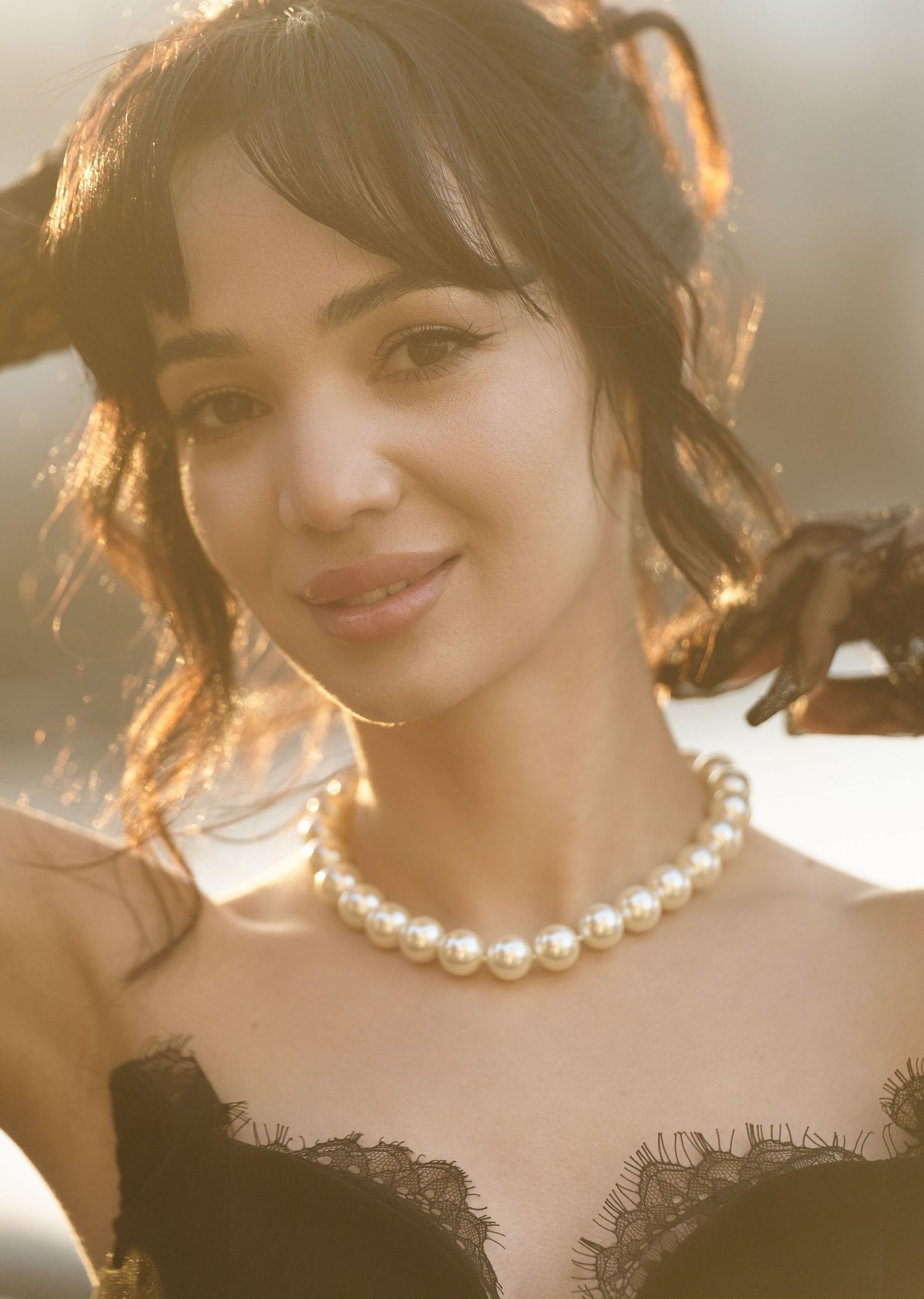 Woman in black lace dress with pearl necklace poses in warm golden sunlight.