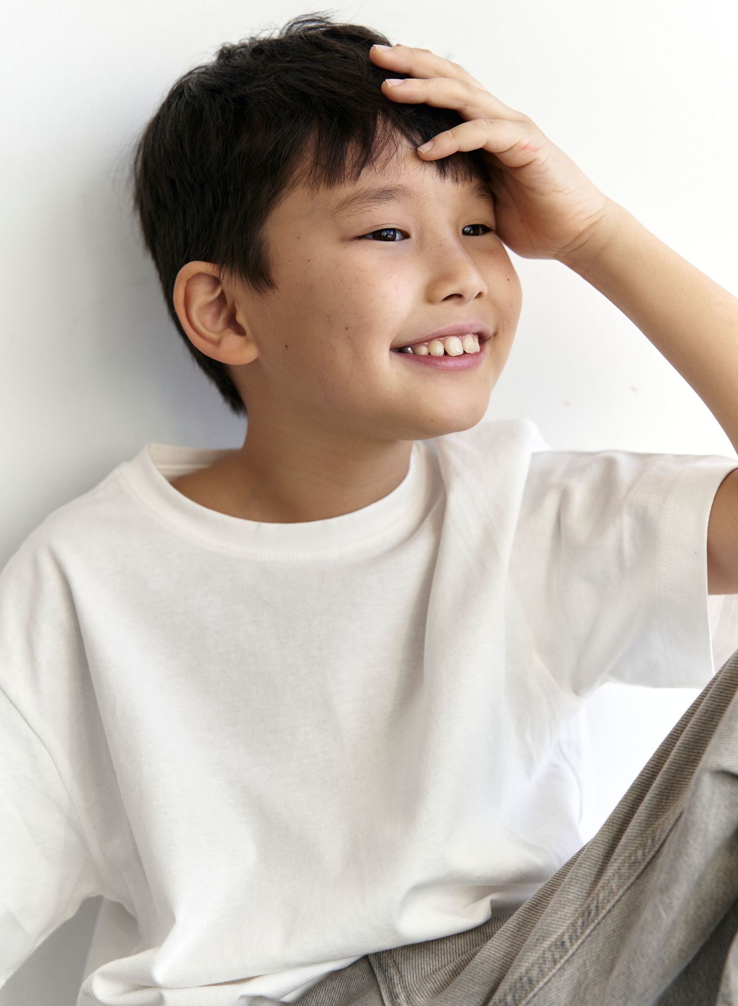 Smiling young boy in white t-shirt leans against wall with hand on forehead.