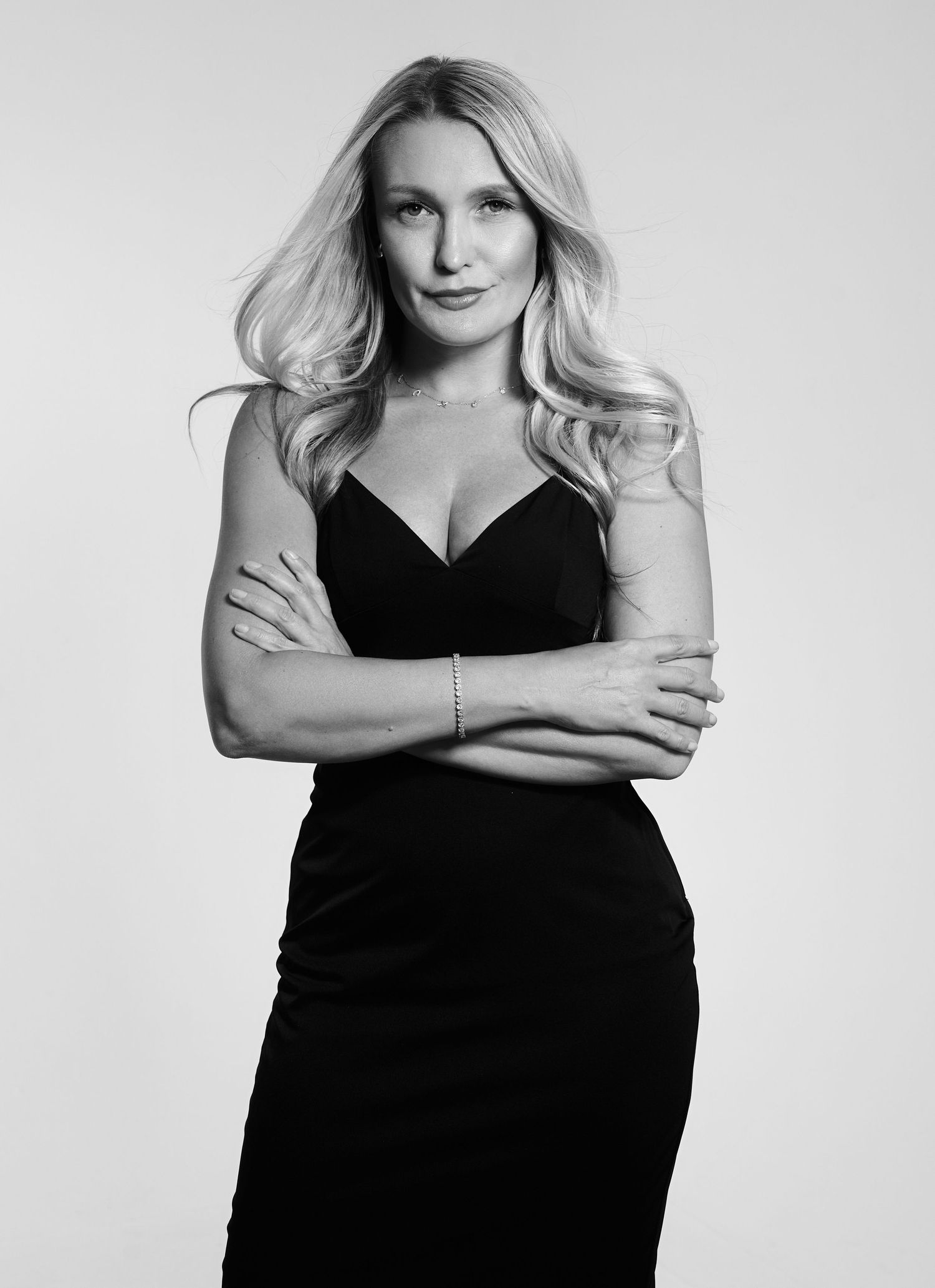 Blonde woman in black spaghetti strap dress with arms crossed in black and white studio portrait.
