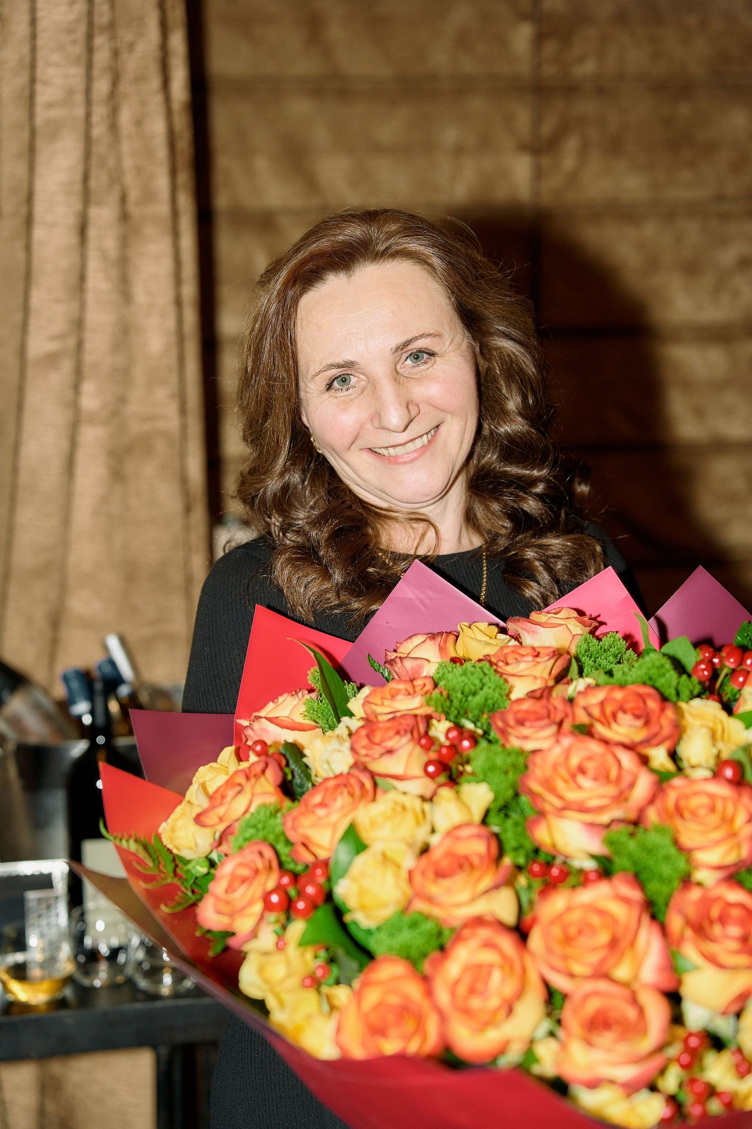 Smiling woman with curly hair holds a large bouquet of orange and yellow roses at a celebration.