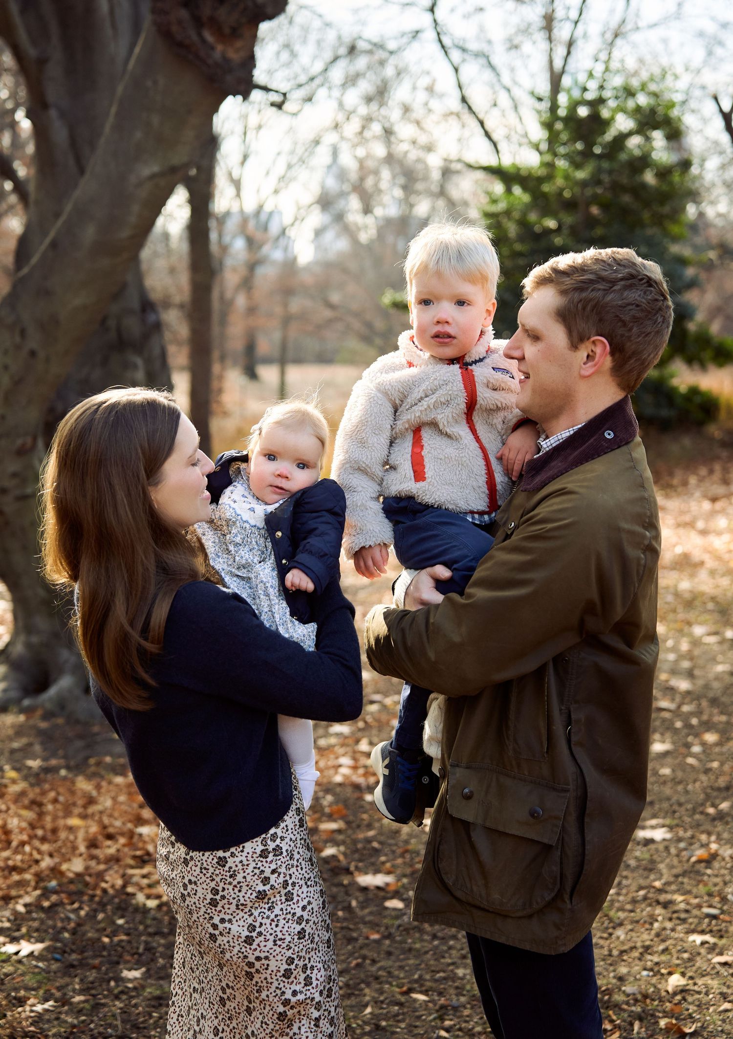Family portrait of parents holding two young children outdoors in a park during autumn.