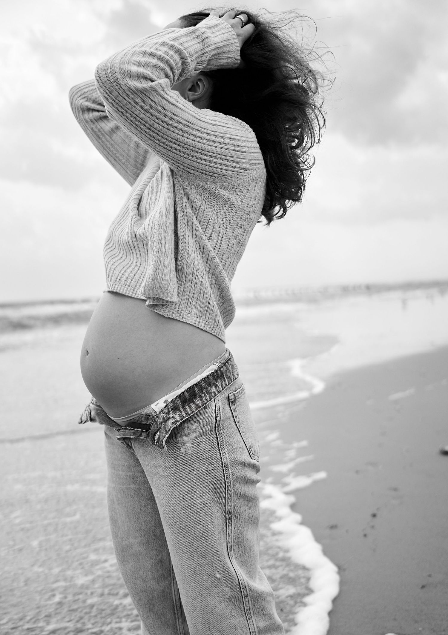 Black and white beach maternity photo of pregnant woman in cropped sweater and jeans by ocean waves.