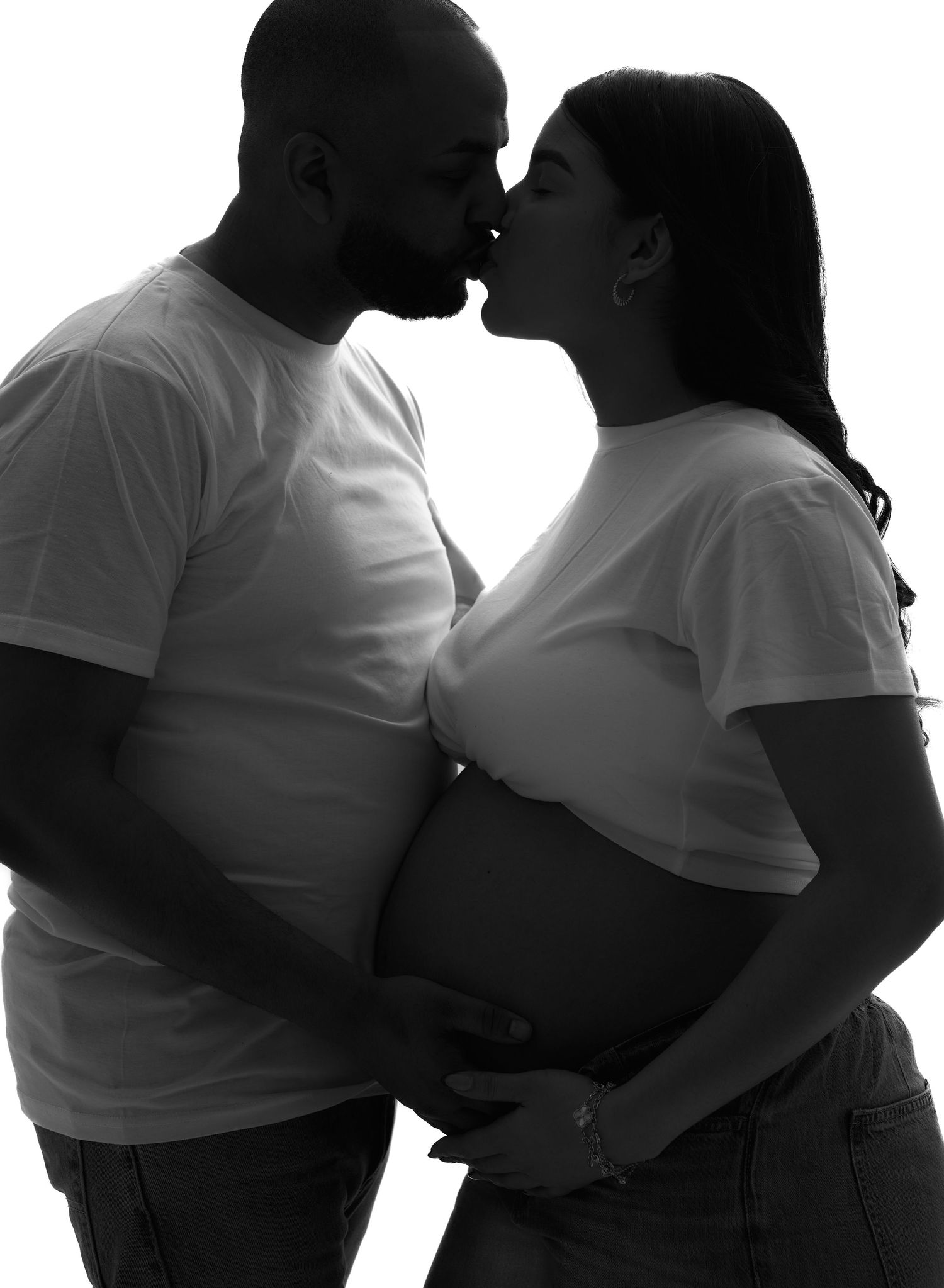Black and white maternity photo of a couple kissing while cradling pregnant belly in a studio.