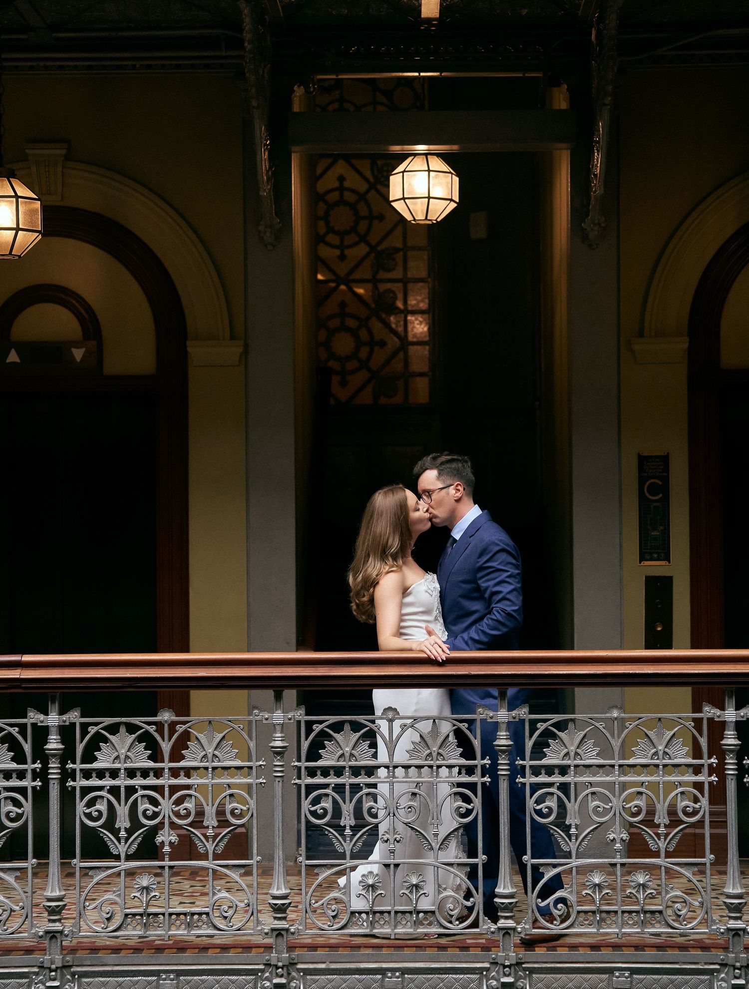 Romantic couple sharing a kiss on decorative iron balcony inside a warmly lit historic venue.